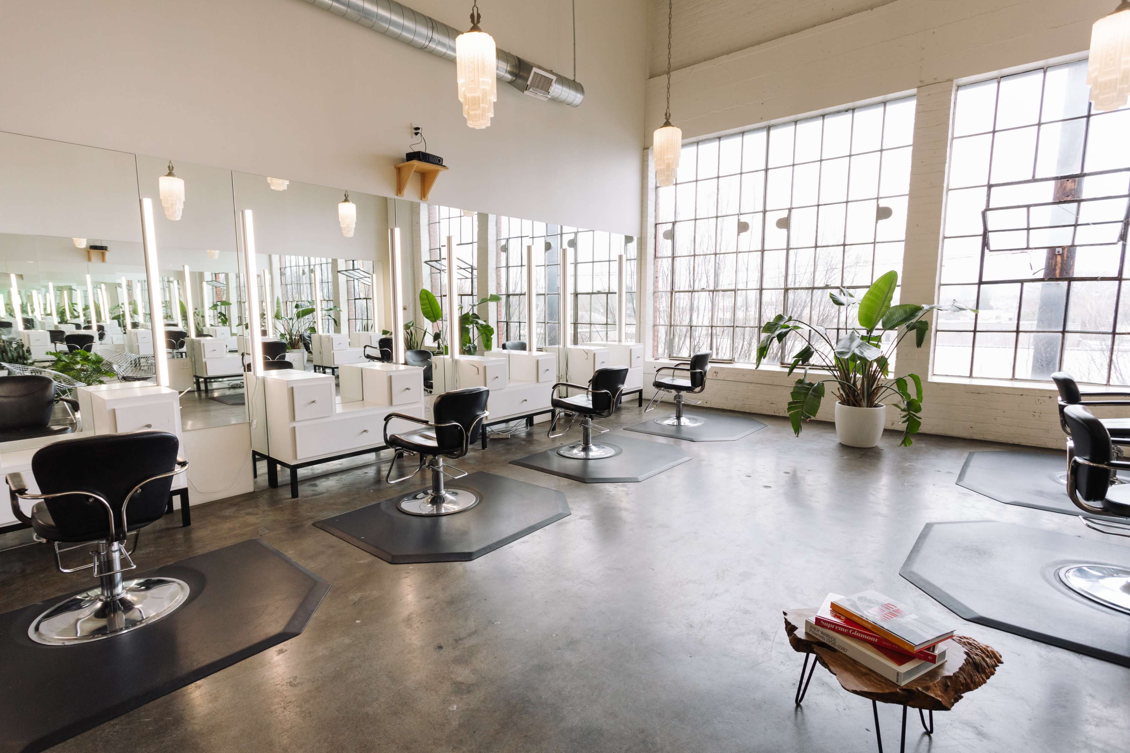 A spacious hair salon with several styling stations, large windows, and potted plants.