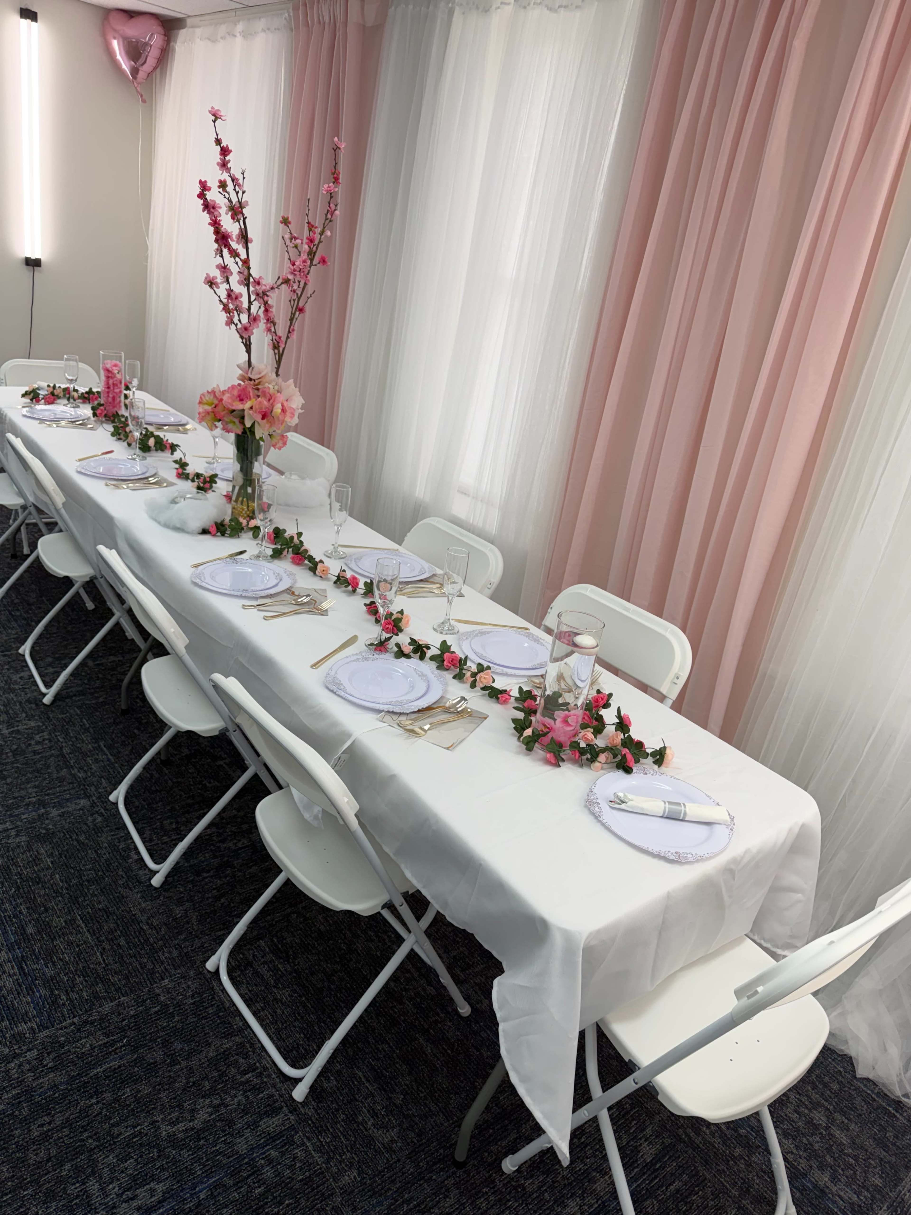 A long table is set for a gathering, adorned with white linens, flowers, and place settings, with pink curtains and decorations in the background.