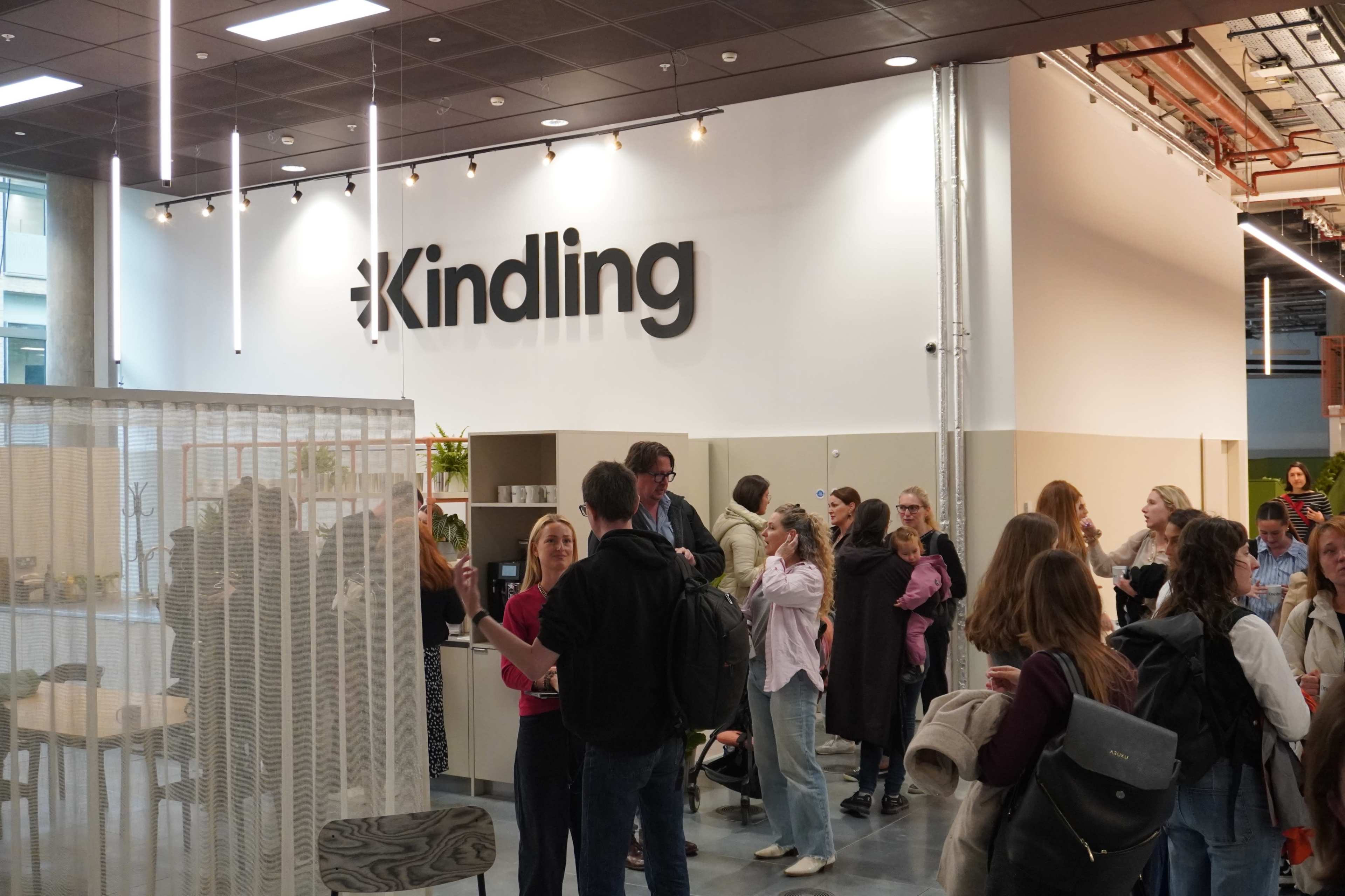 The image shows a modern indoor space with a prominent sign that reads "Kindling," and a group of people gathered near a reception area.