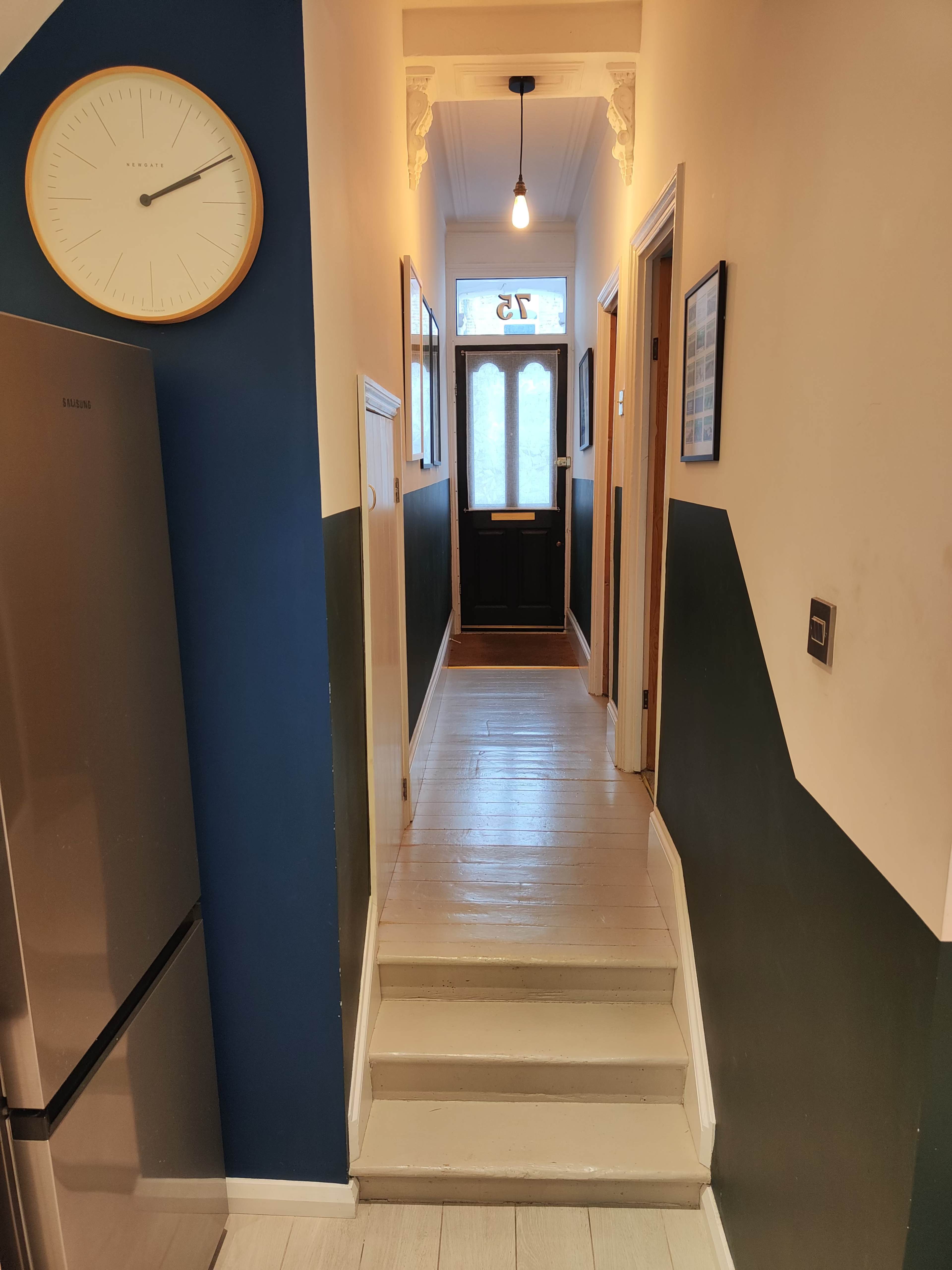 A hallway with a wooden floor, featuring a clock on the wall, a refrigerator to the left, and a front door at the end of the corridor.