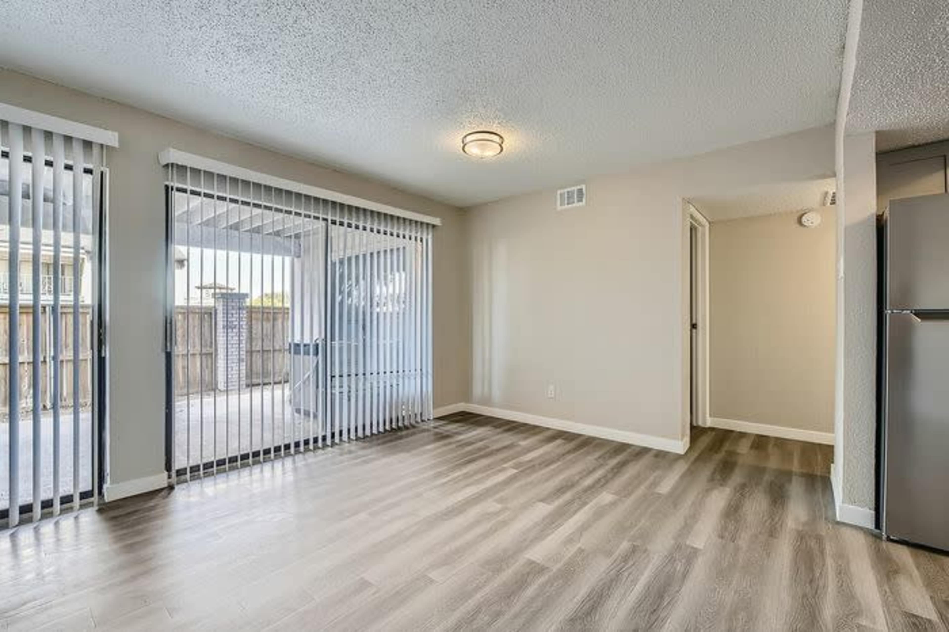 The interior of a room features beige walls, a ceiling light, and sliding glass doors leading to an outdoor patio area, with laminate flooring throughout.