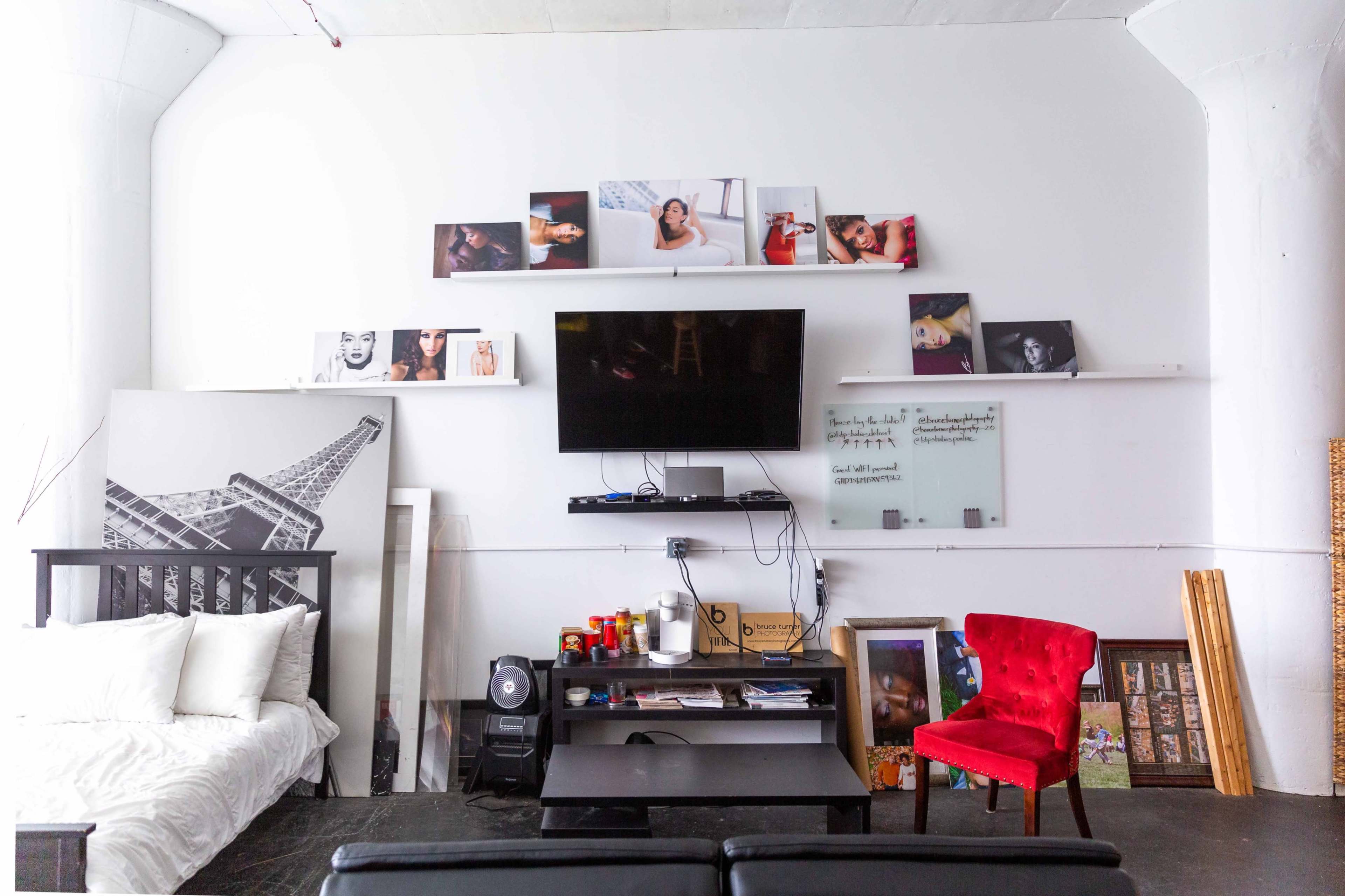 The image shows a modern living space featuring a bed, a coffee table, a red chair, and various artworks and photographs displayed on the wall above a television.