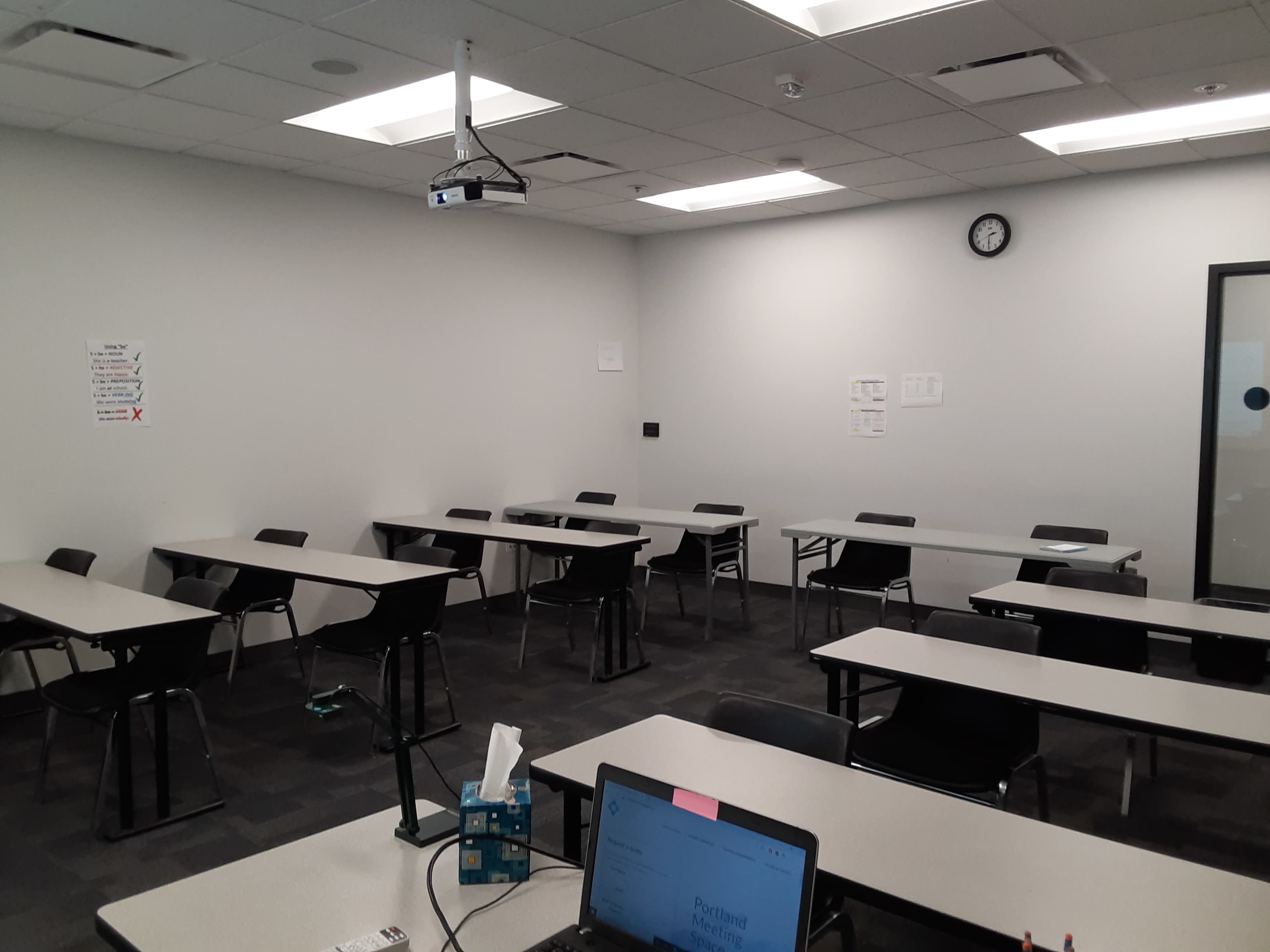 A classroom features several tables and chairs arranged in rows, with a projector mounted on the ceiling and a clock on the wall.