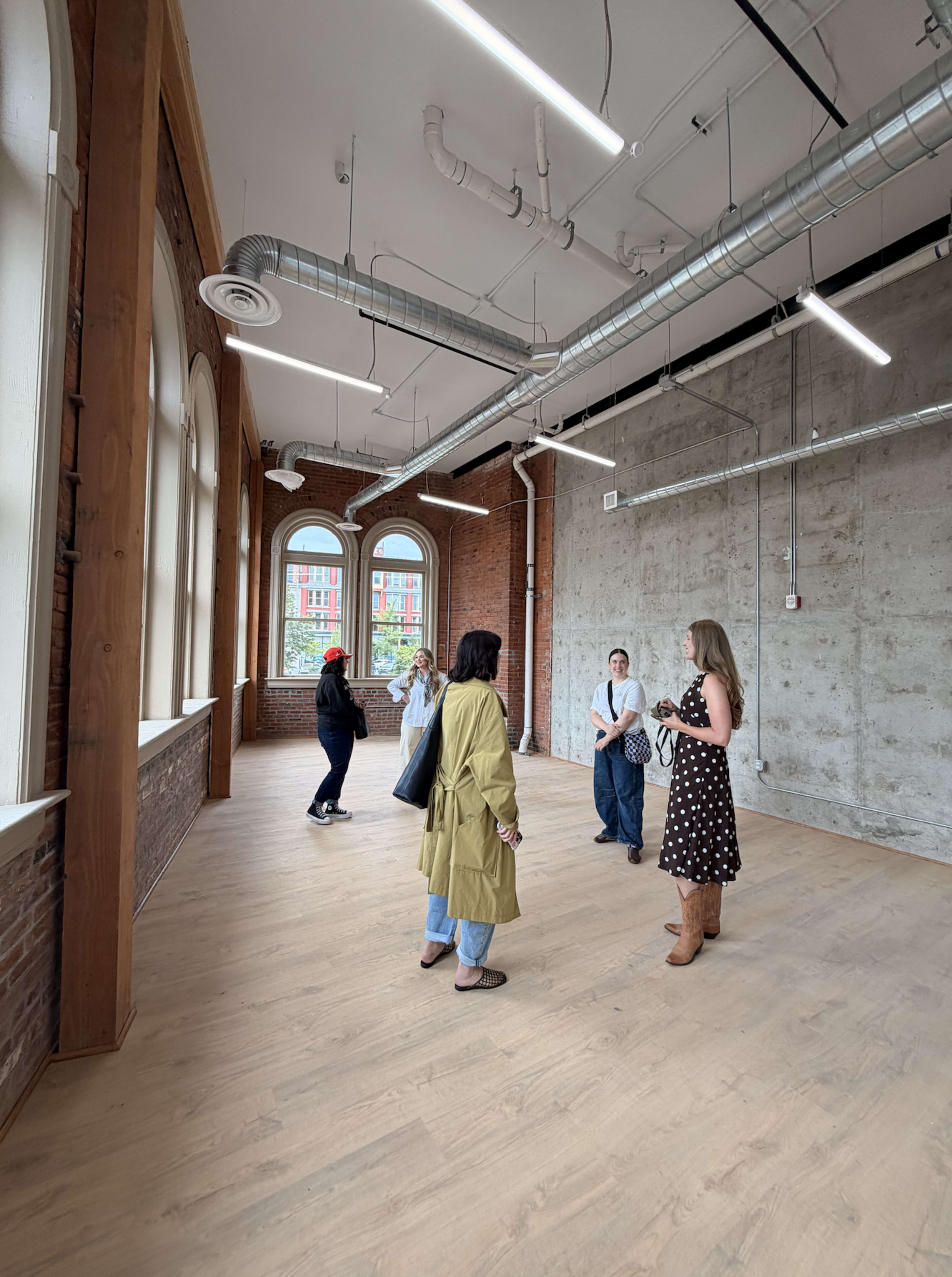 The image shows a group of five individuals interacting in a spacious room with large windows, exposed brick walls, and wooden flooring.