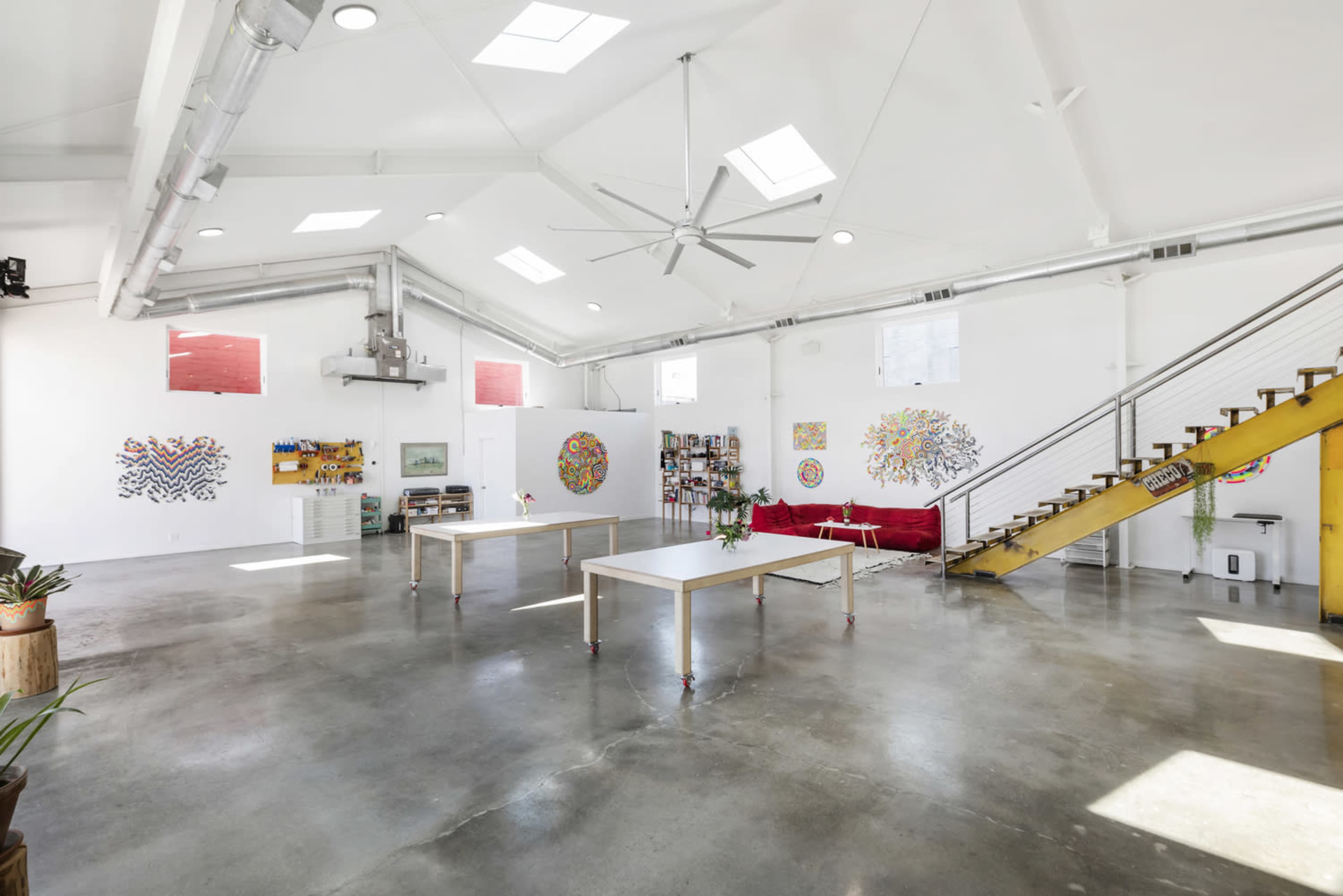 The image shows an open, brightly lit loft space with concrete floors, high ceilings, and colorful artwork scattered on the white walls, along with a red sofa and a staircase leading to an upper level.