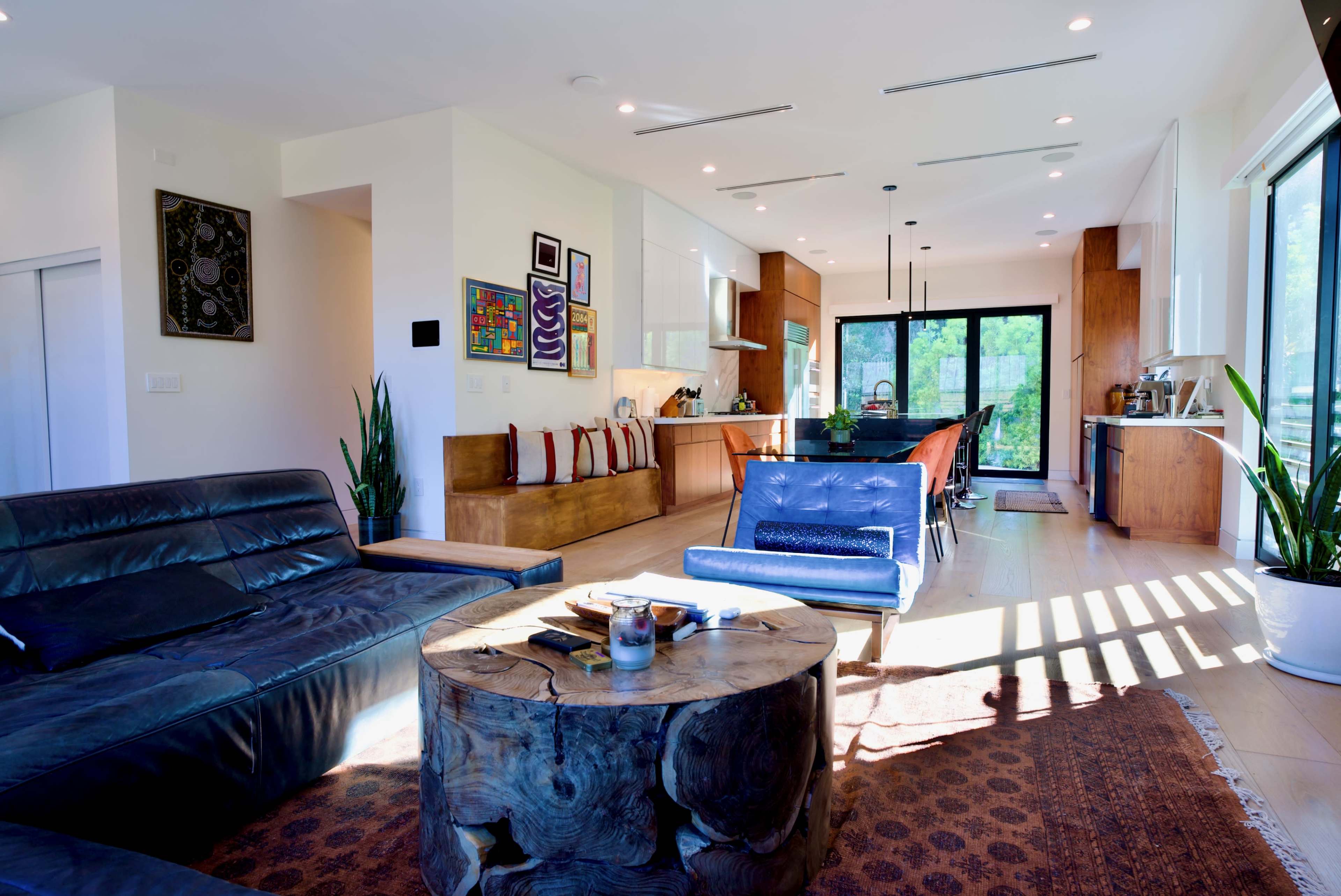 The image shows a modern living room with a combination of seating areas, a wooden dining table, and large windows allowing natural light to fill the space.