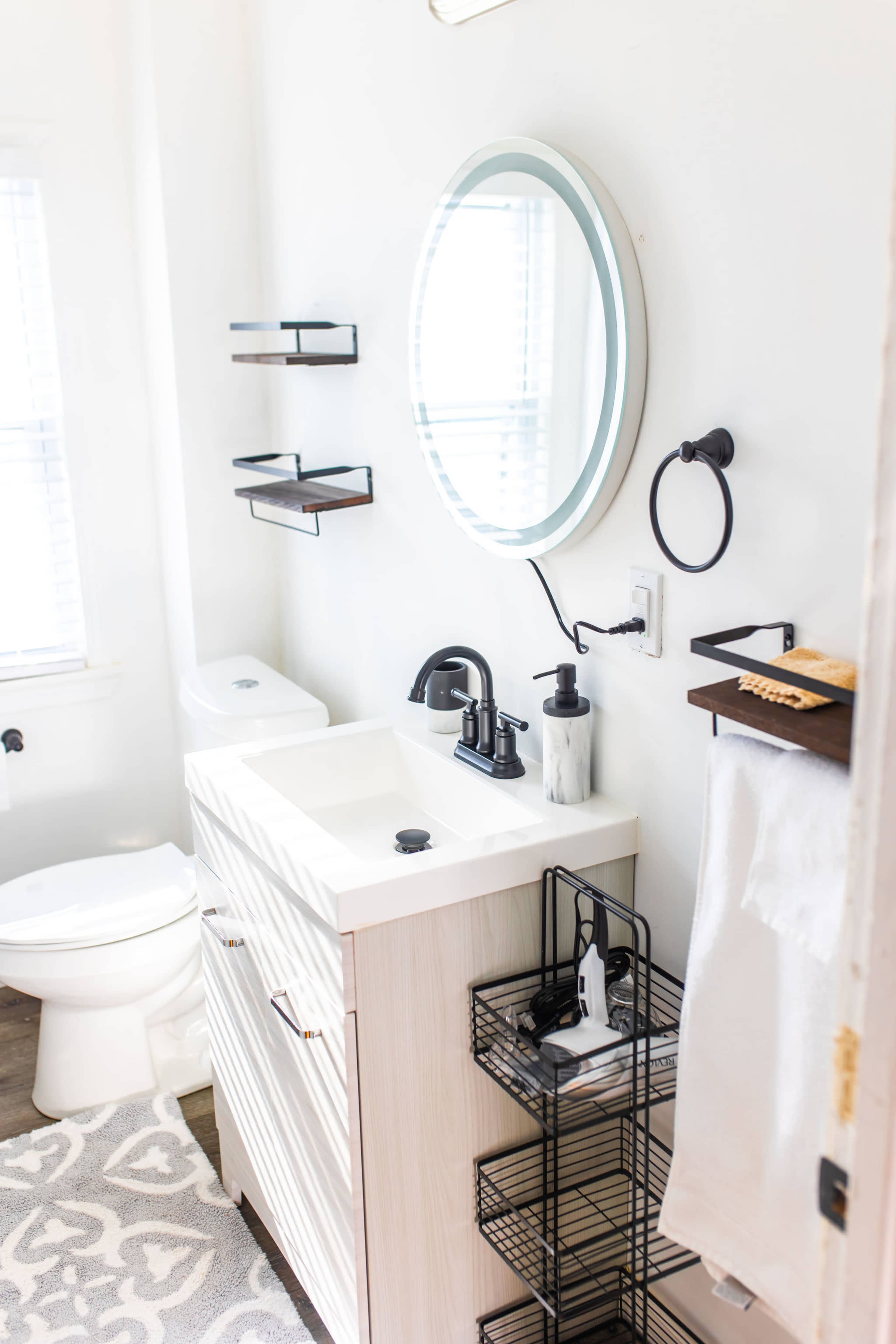 A modern bathroom featuring a round mirror, sink with a black faucet, storage shelves, and a towel rack on the wall.