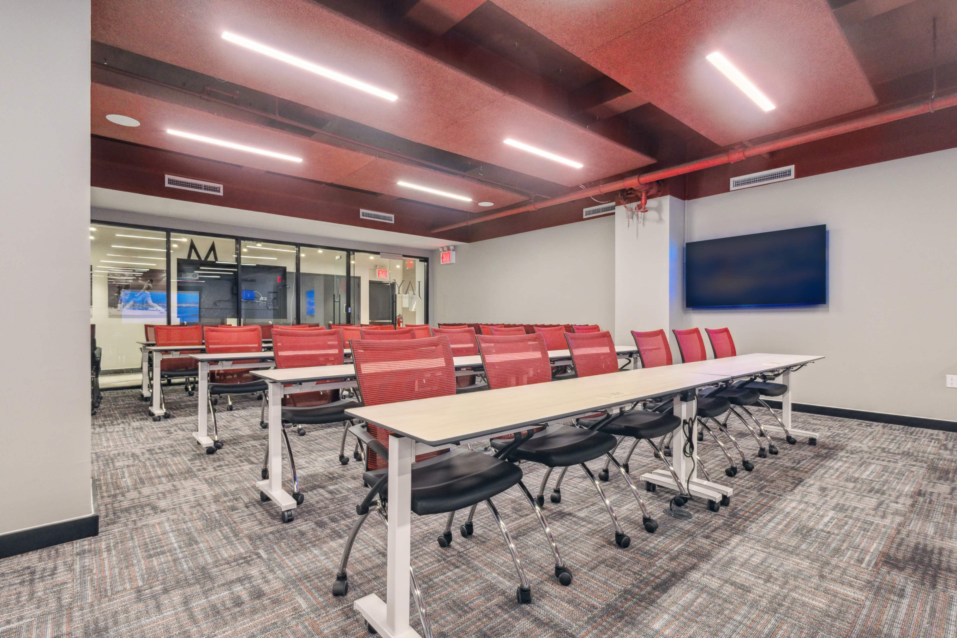 A modern conference room features a long table with rolling chairs arranged neatly, illuminated by overhead lights and equipped with a wall-mounted screen.