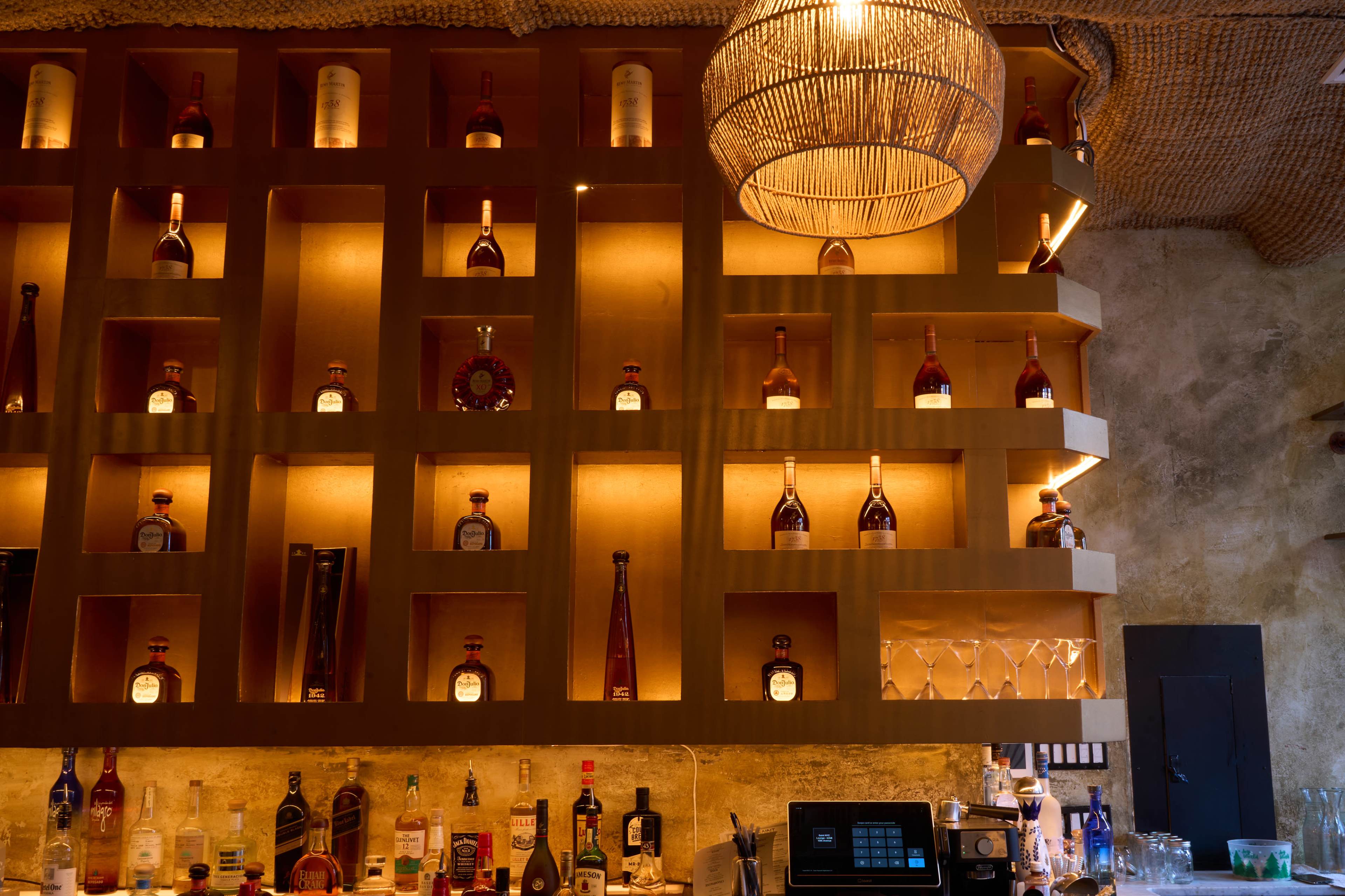 The image features a well-lit bar with a structured display of various liquor bottles arranged in a wooden shelf.
