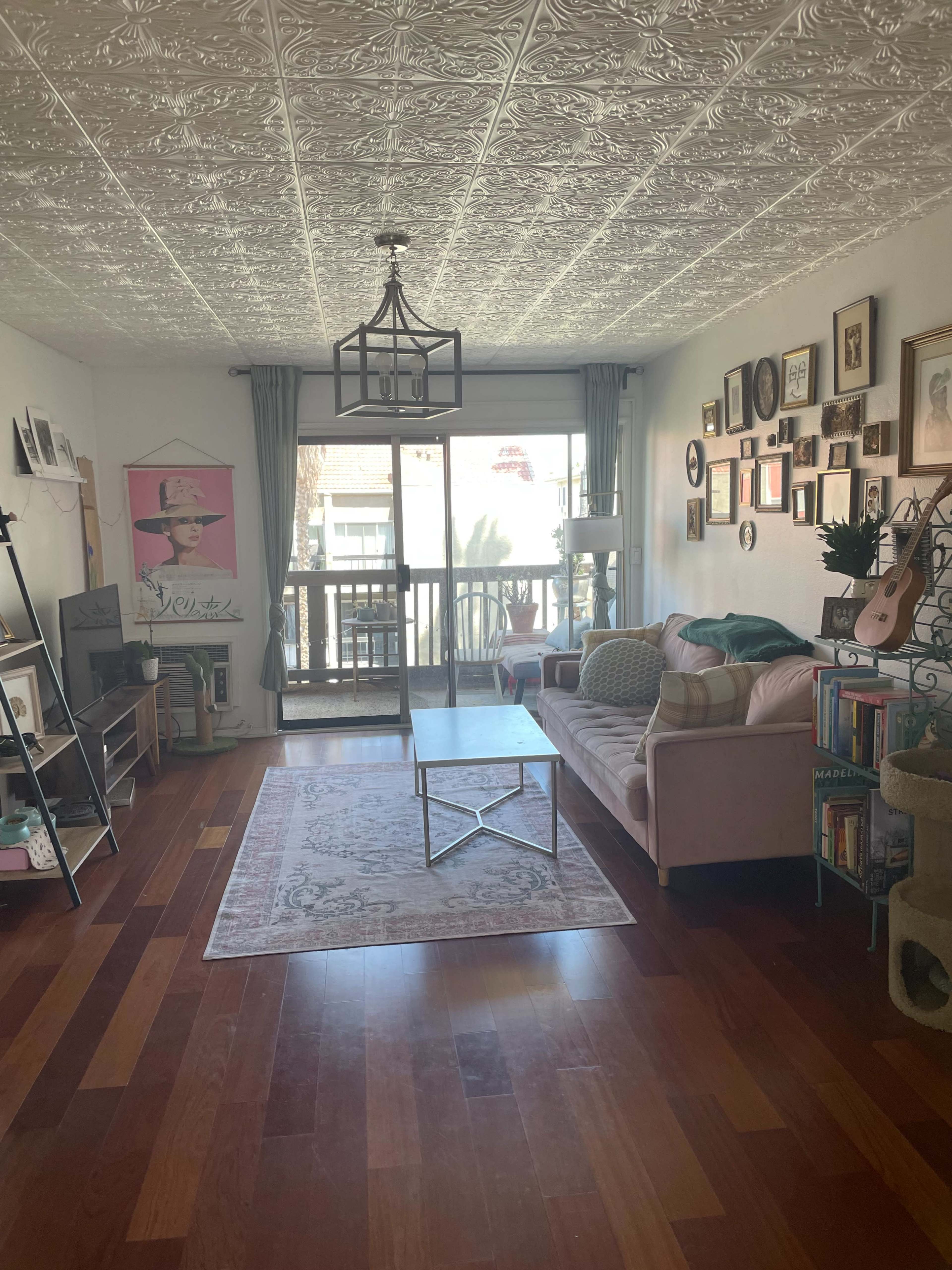 The living room features a pink sofa, a coffee table, and a large window with sliding glass doors leading to a balcony, along with various wall art and shelves.