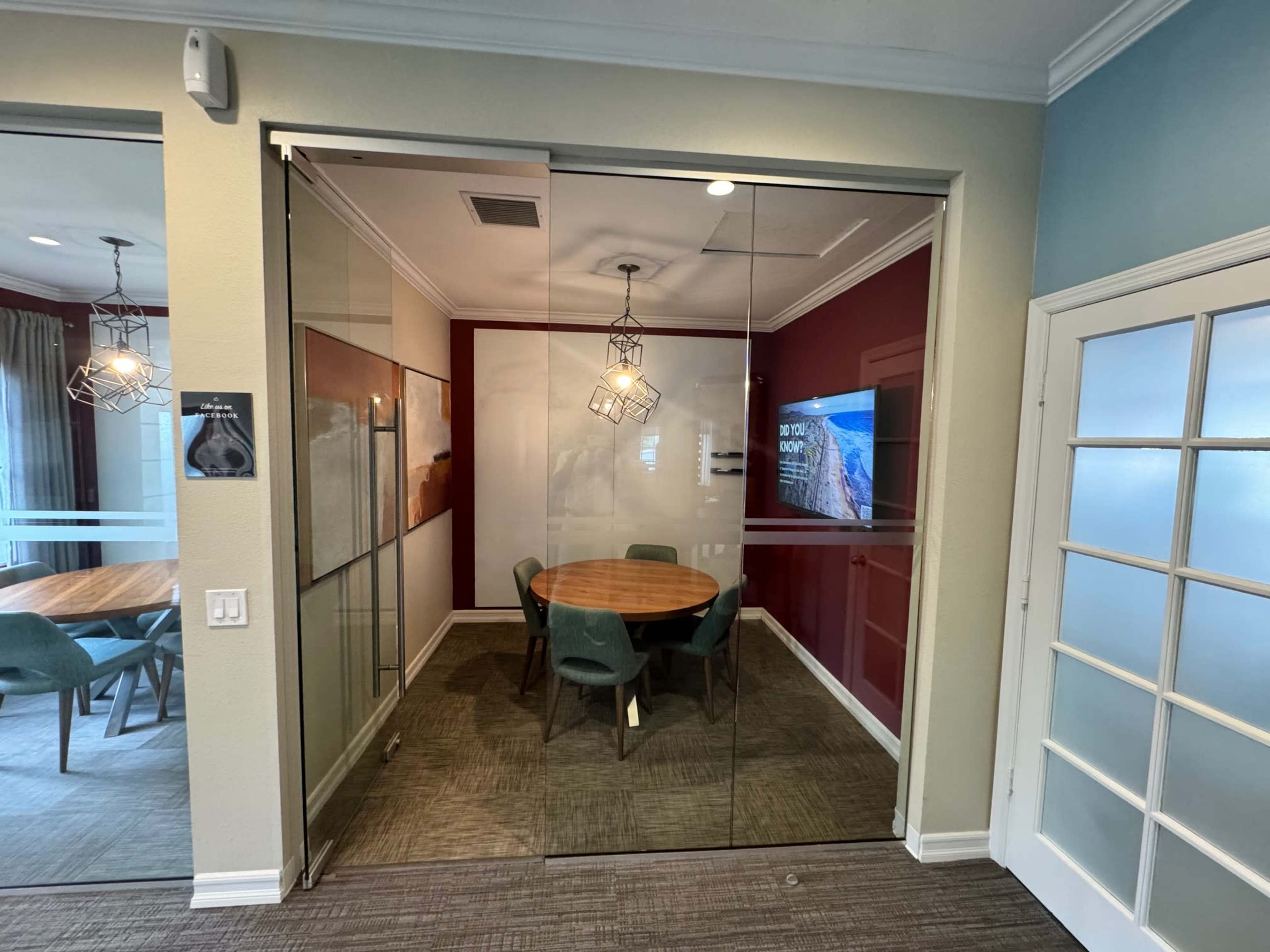 A glass-walled conference room features a round wooden table surrounded by chairs and a large screen on the wall.