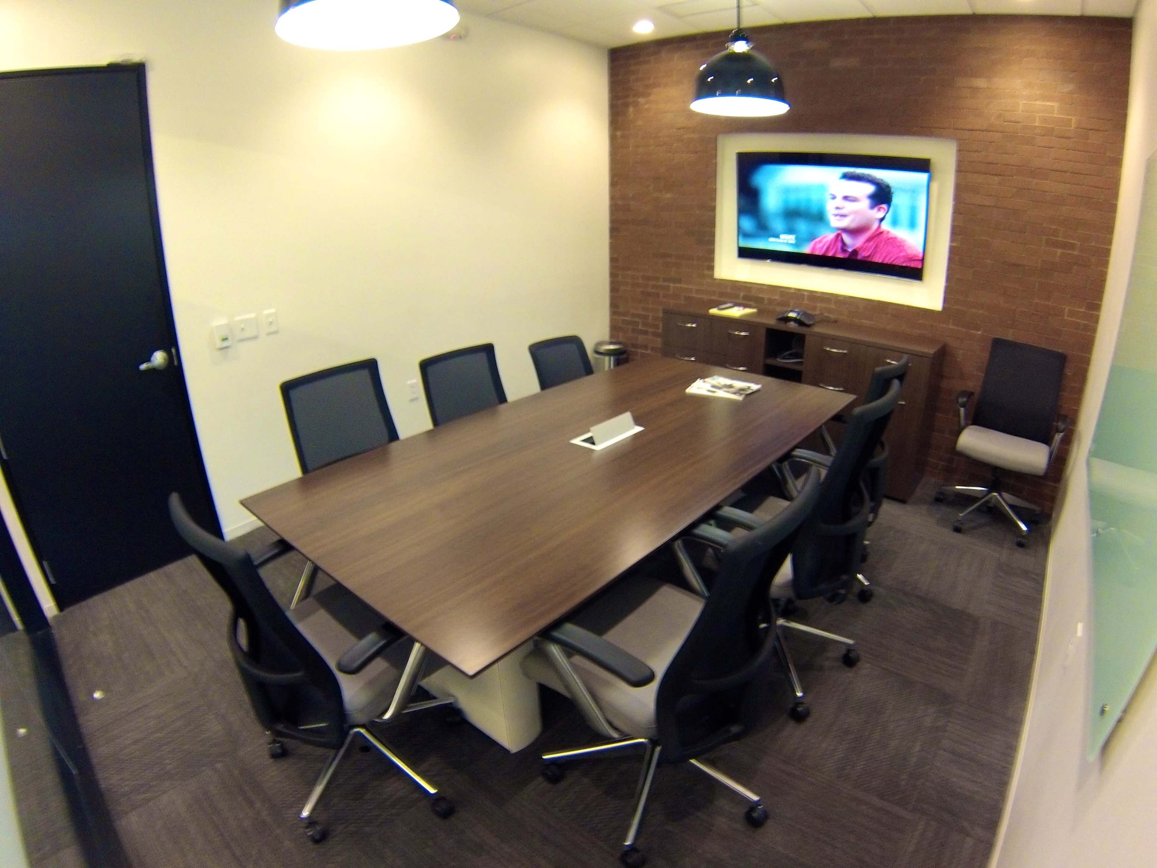 A conference room features a long wooden table surrounded by eight chairs, with a television mounted on the wall displaying a presentation.
