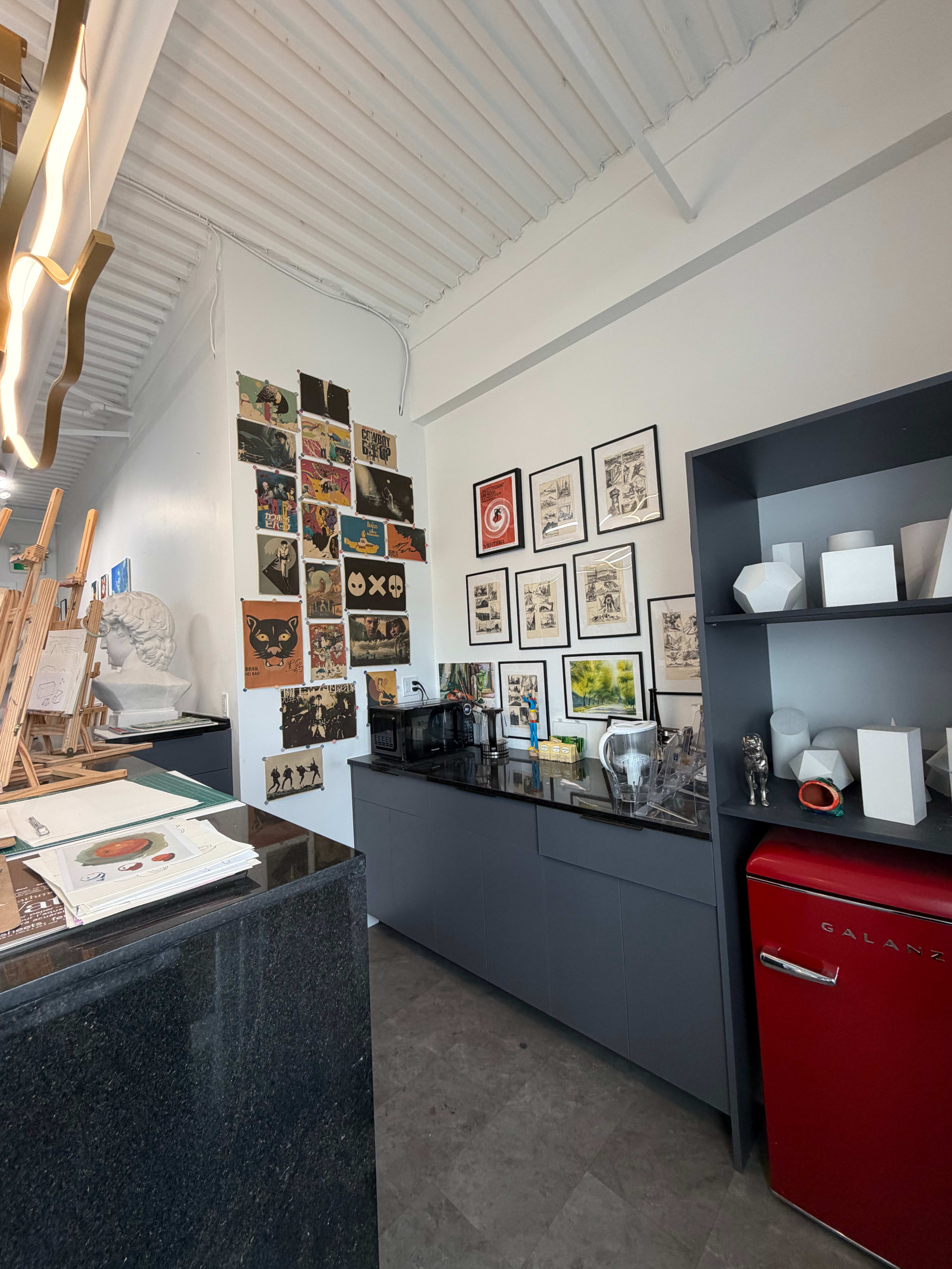The image shows a modern café interior featuring a dark countertop and a gallery wall adorned with various framed artworks, alongside a red refrigerator and a shelf displaying decorative items.