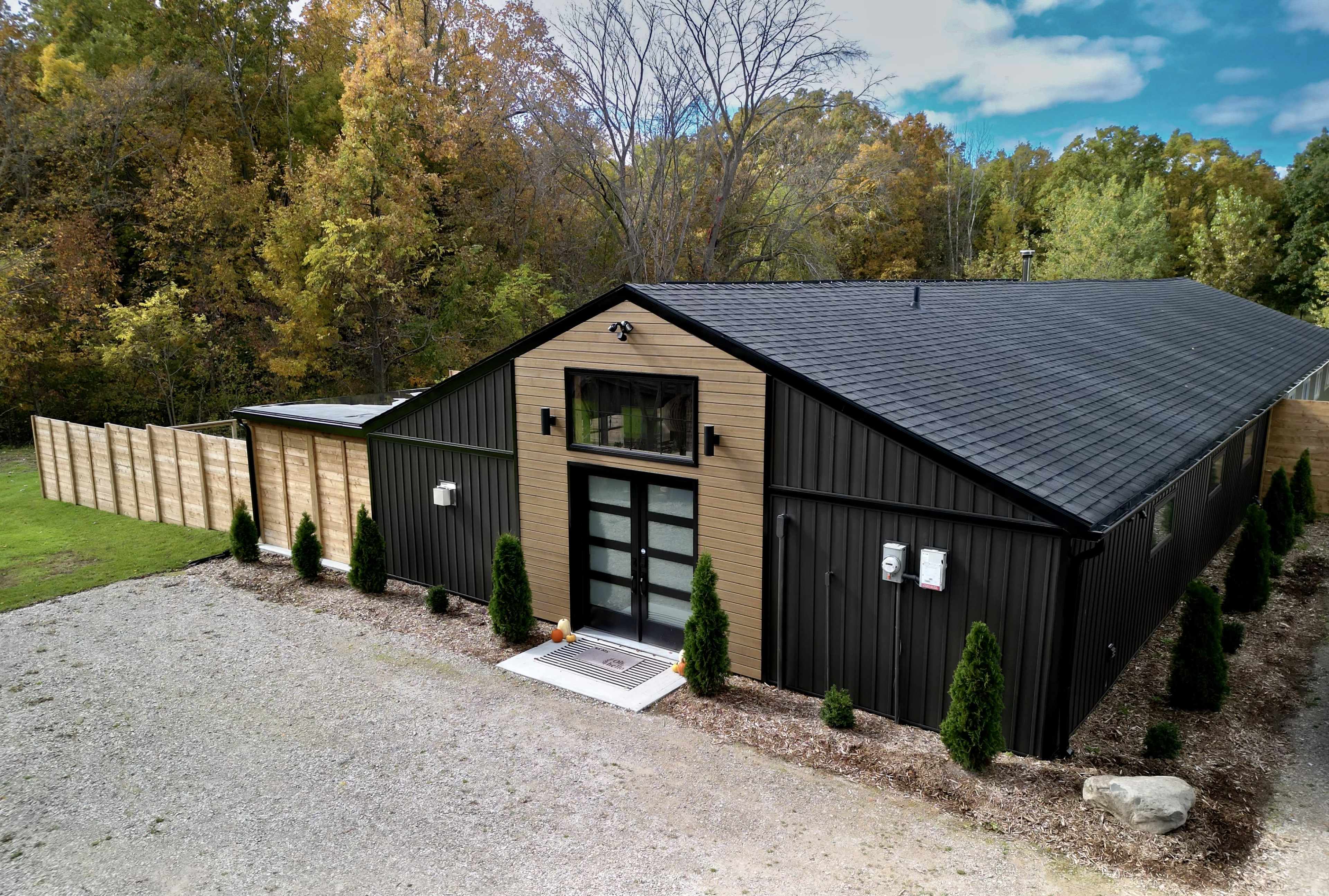 A modern, dark-colored building with a large glass entrance is surrounded by neatly trimmed shrubs and a gravel driveway, set against a backdrop of trees with autumn foliage.