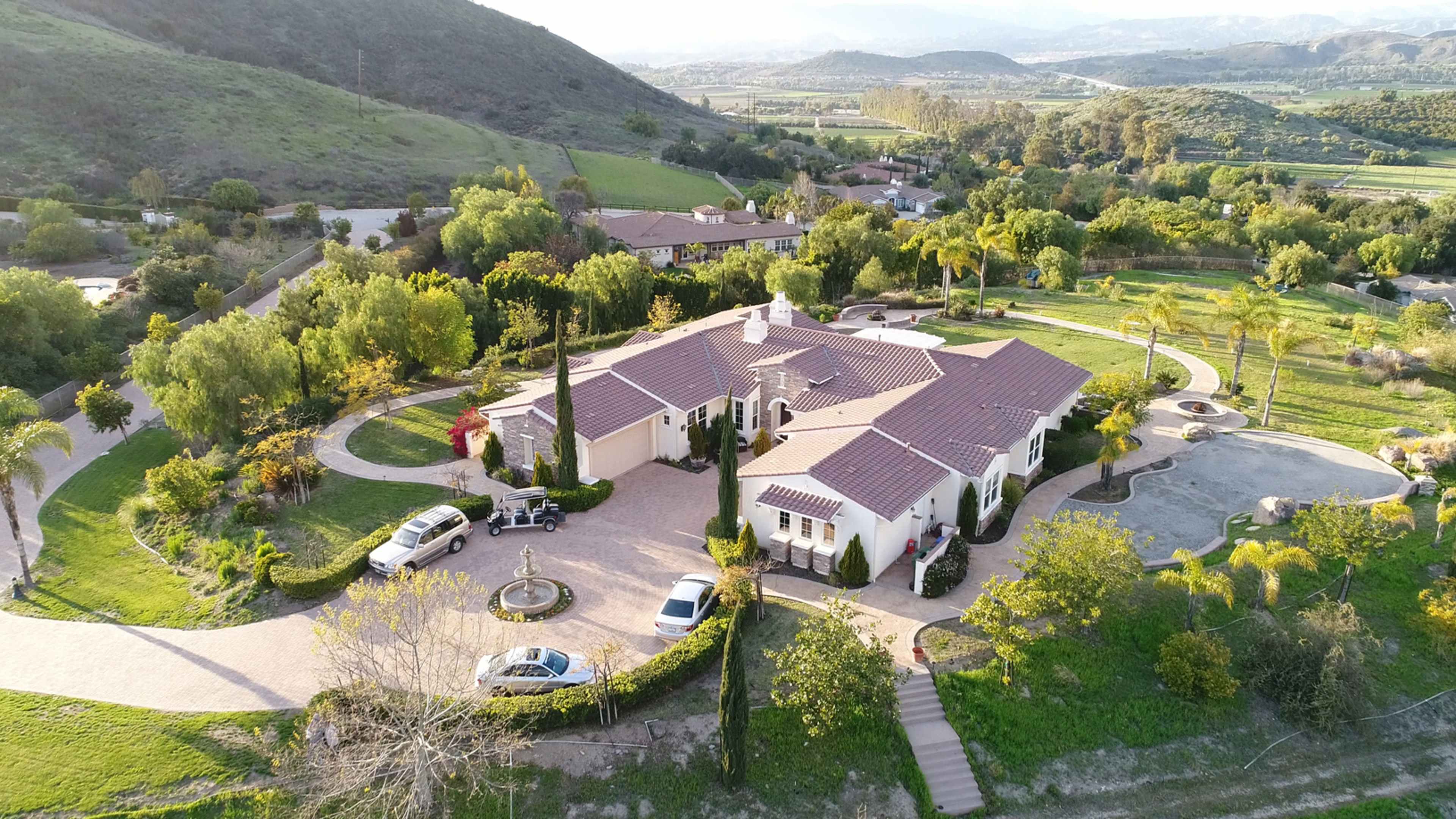 A large, sprawling residential property surrounded by greenery and hills, with multiple vehicles parked in the driveway.
