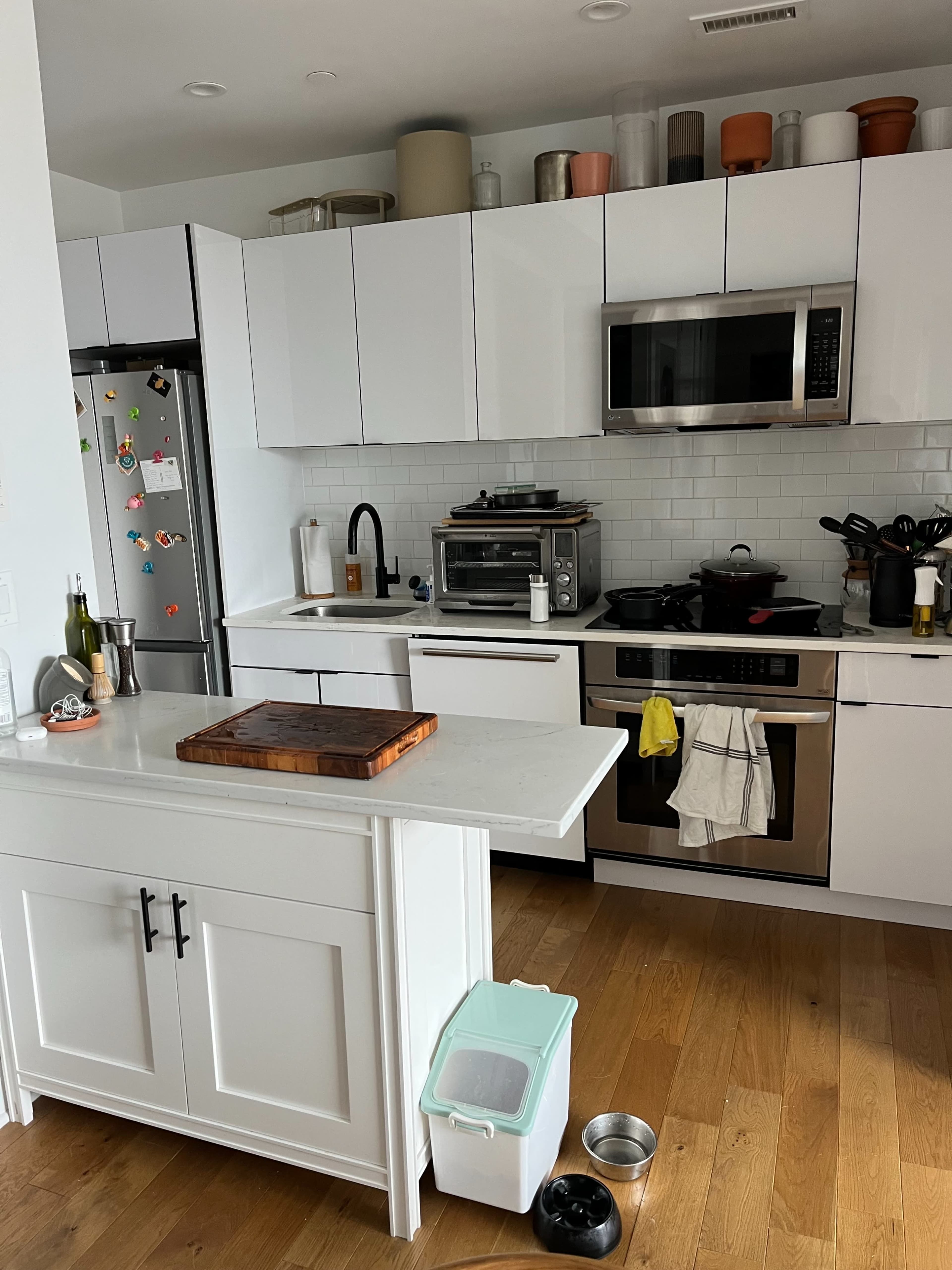 The image shows a modern kitchen with white cabinets, stainless steel appliances, and a wooden floor, featuring a central island and various kitchen items on the countertops.