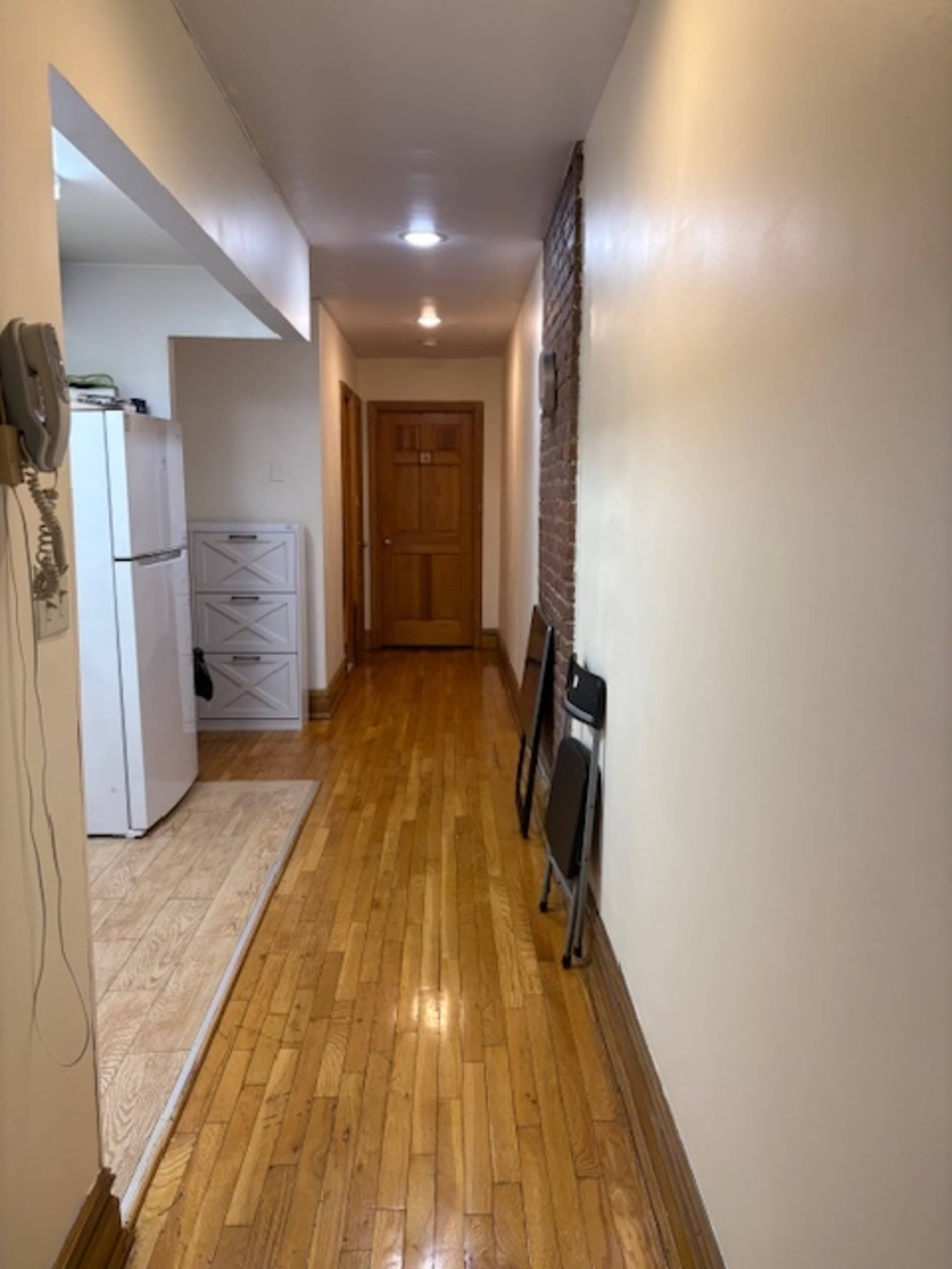 The image shows a long, narrow hallway with polished wooden floors, a door at the end, and a small kitchen area visible on the left side.