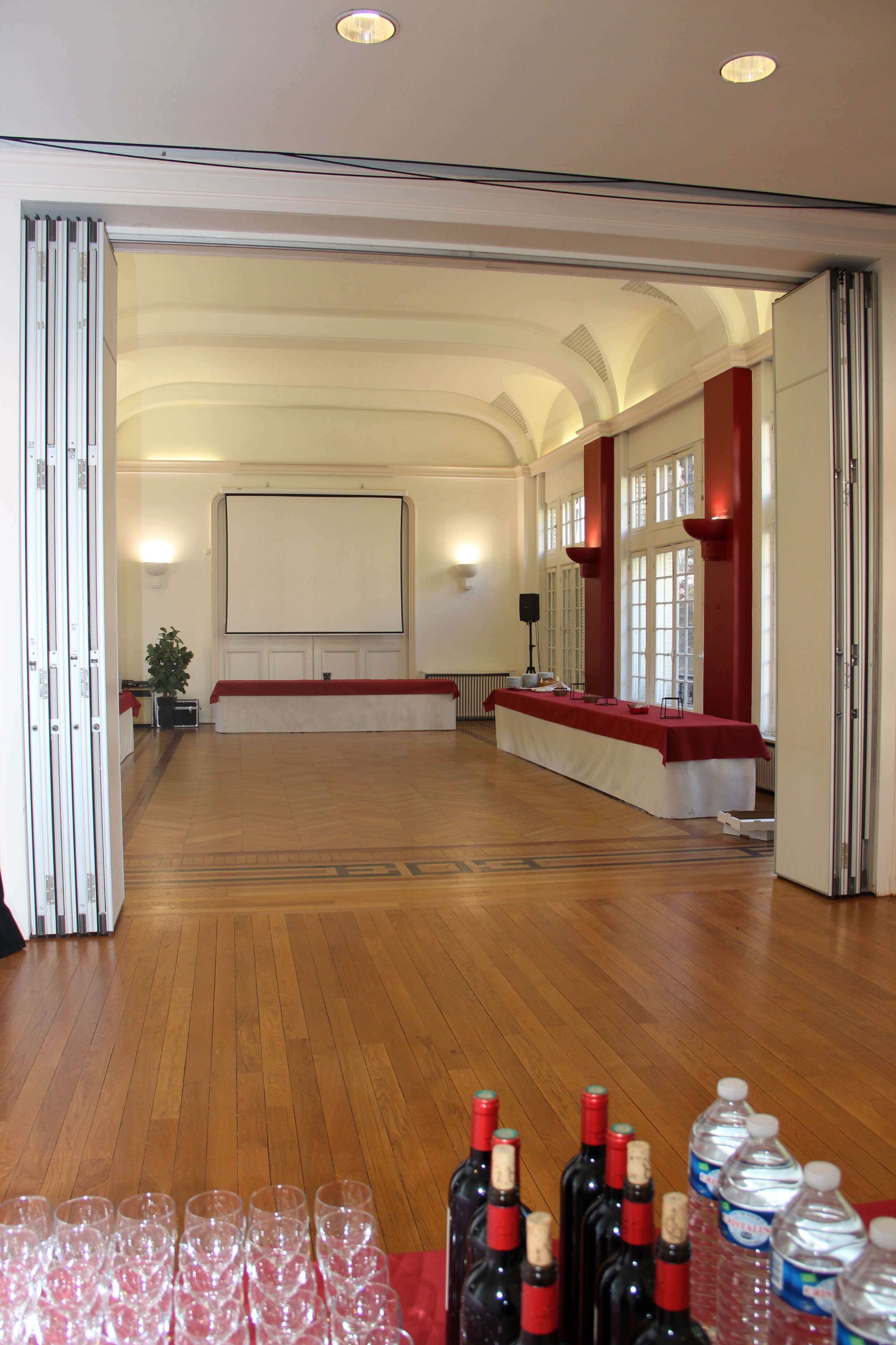 A spacious event room features a large empty stage area with a screen at the front and a table with red tablecloths along the back, accompanied by bottles of red wine and water on a nearby table.