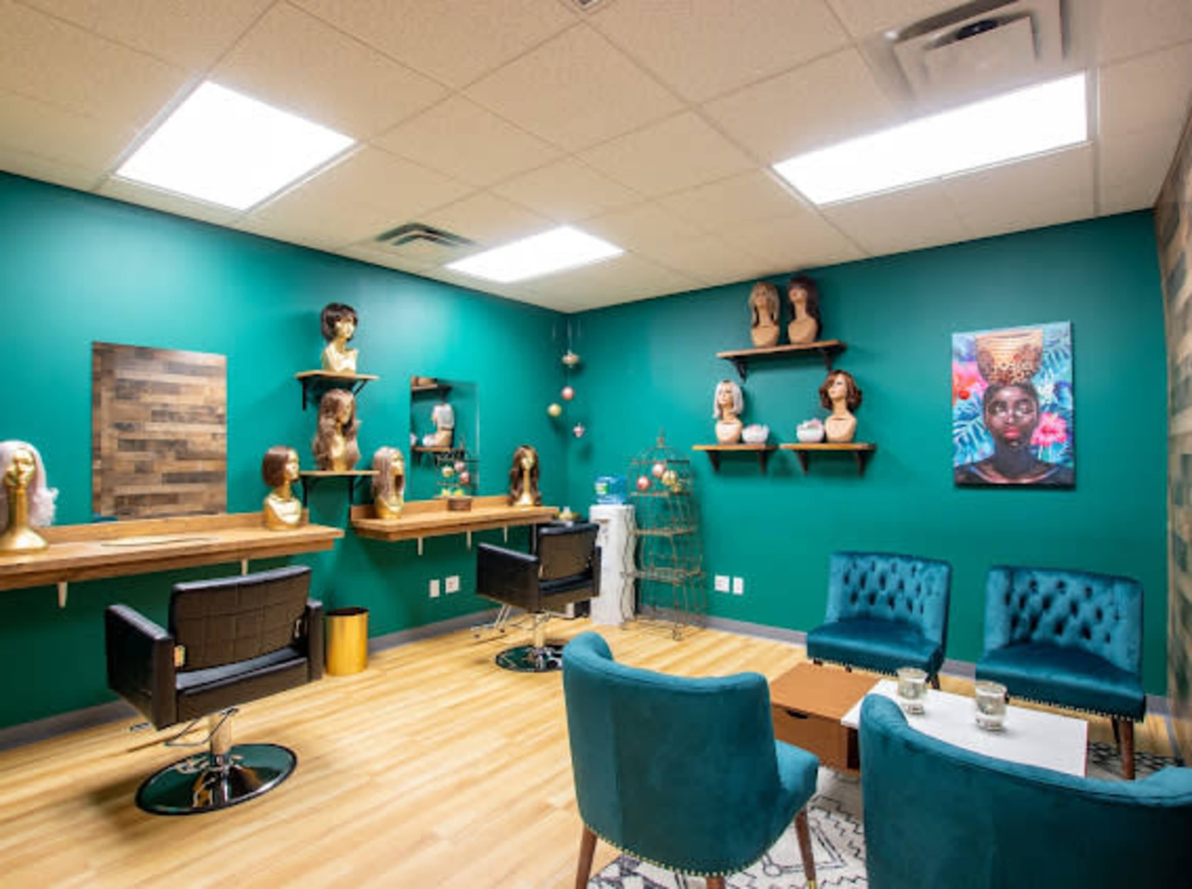 A hair salon interior featuring teal walls, a seating area with teal chairs, a wooden shelf displaying mannequin heads, and artwork on the wall.