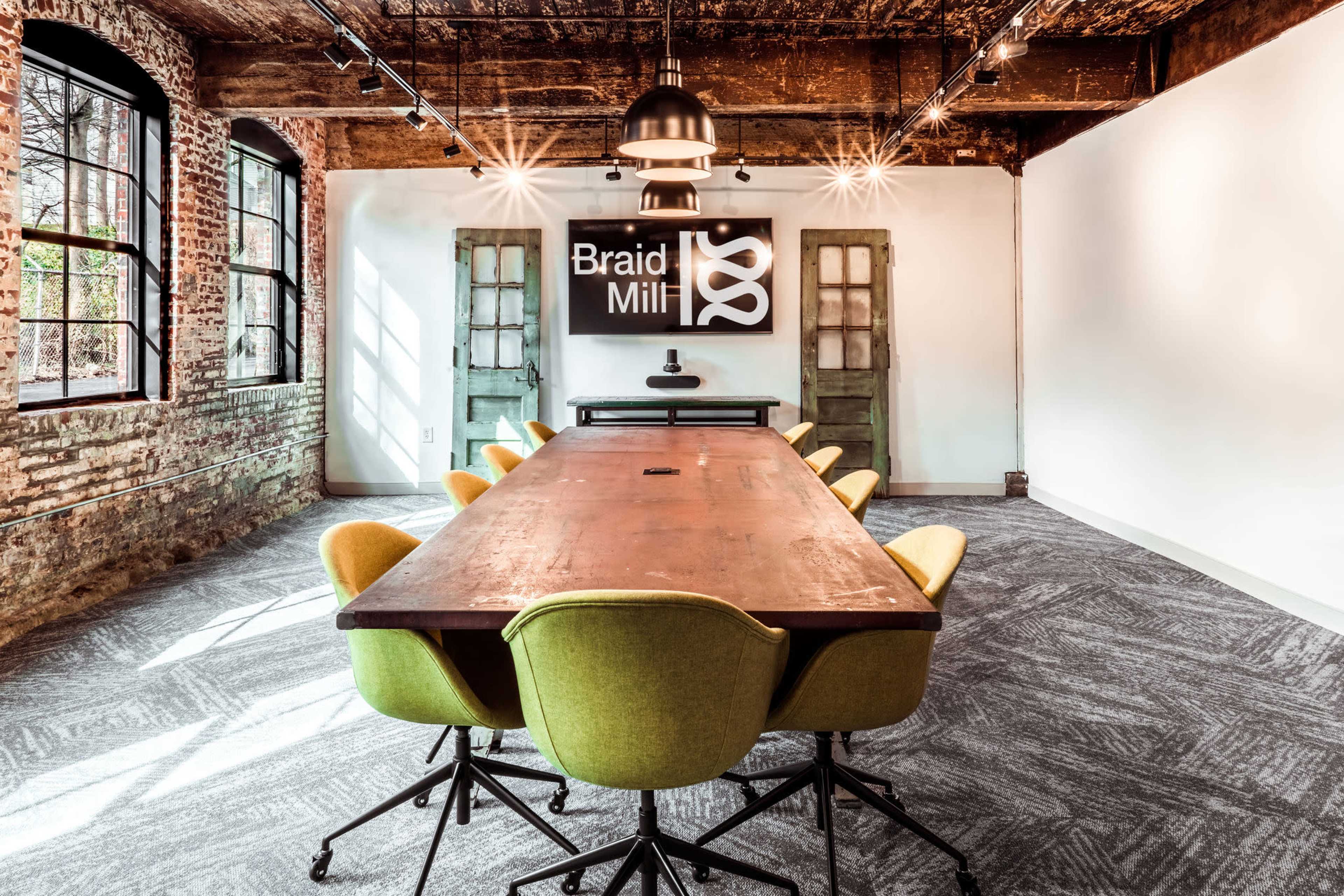 A modern conference room featuring a long wooden table surrounded by yellow chairs, with exposed brick walls and a prominent sign reading "Braid Mill."