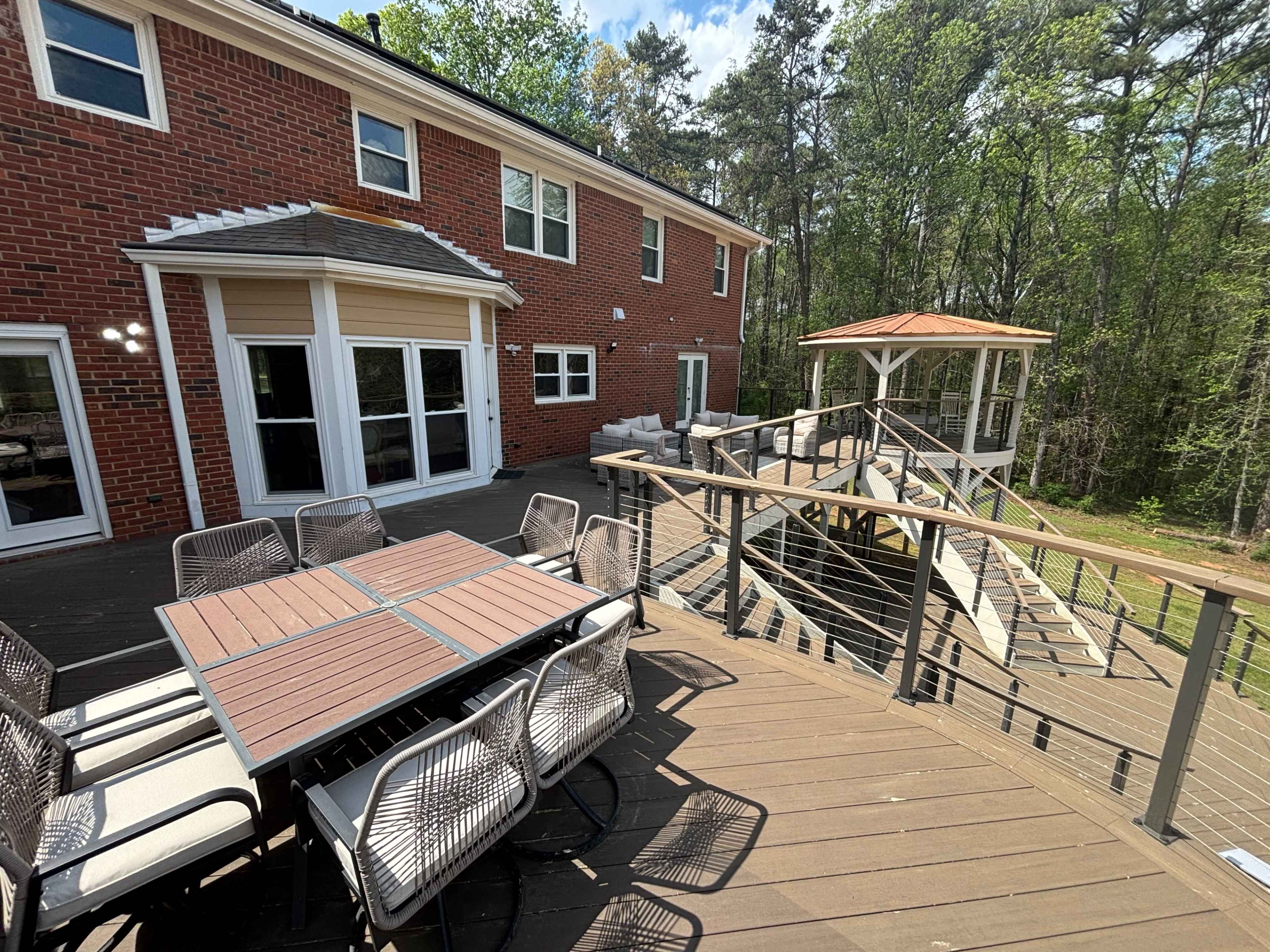 The image shows a backyard patio area with a wooden deck, a large dining table, and a gazebo overlooking a wooded area.