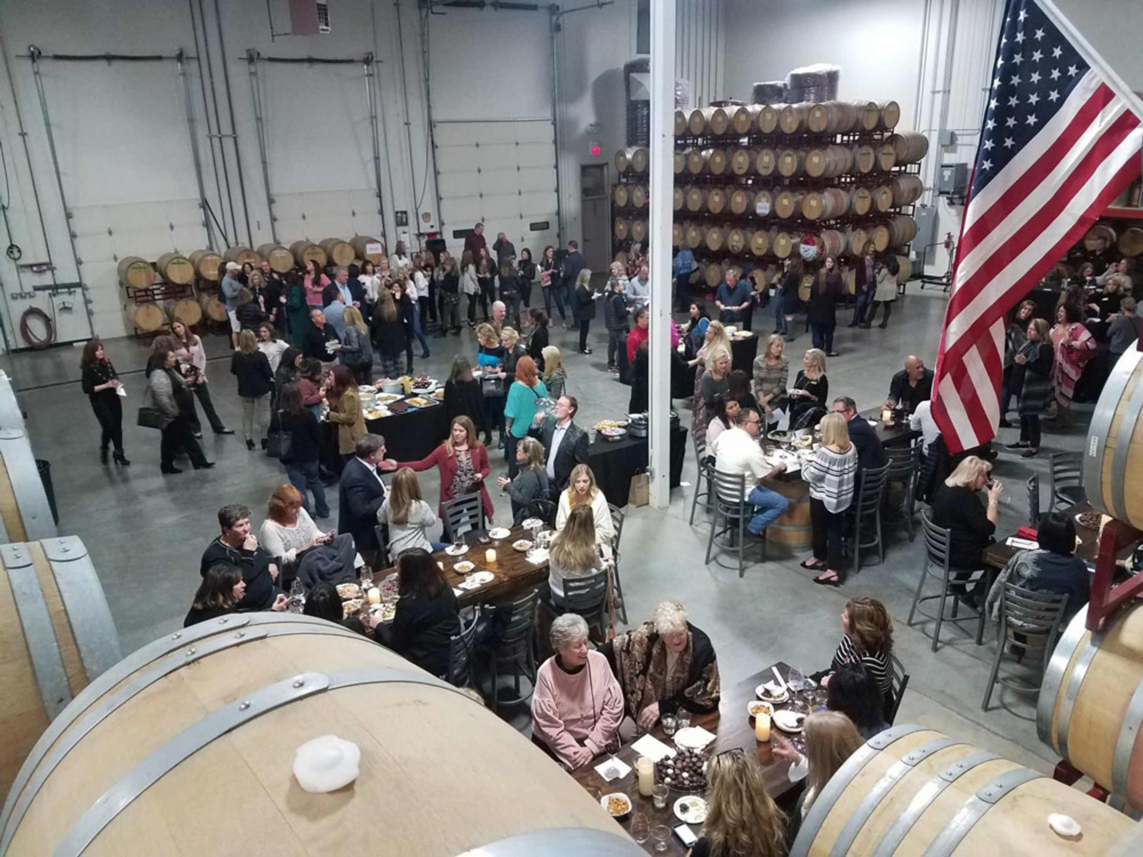 A large gathering of people is held in a warehouse with wine barrels in the background and food platters on tables.