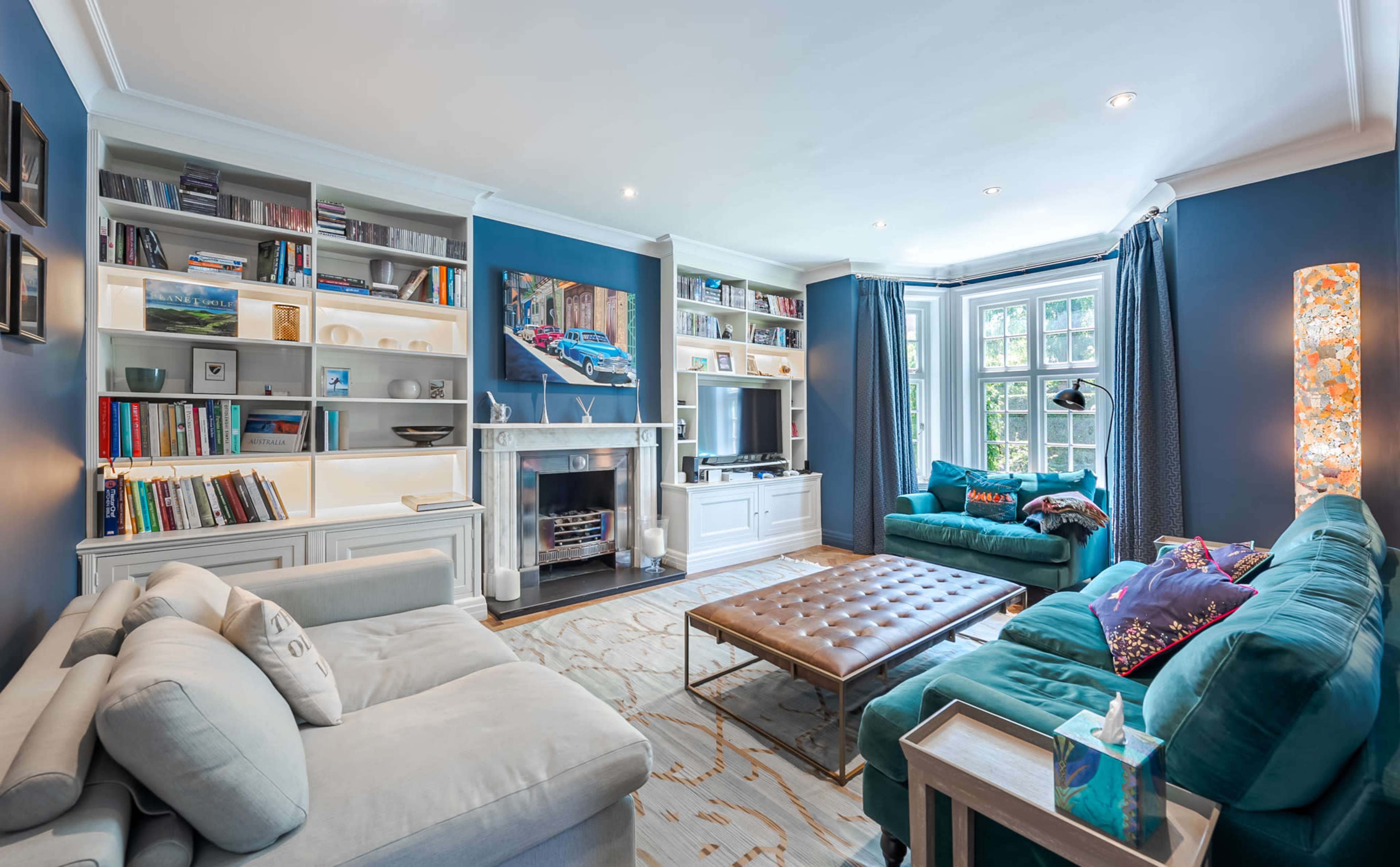 The living room features a blue color scheme with a sectional sofa, two teal armchairs, a bookshelf filled with books, and a large coffee table positioned in the center.