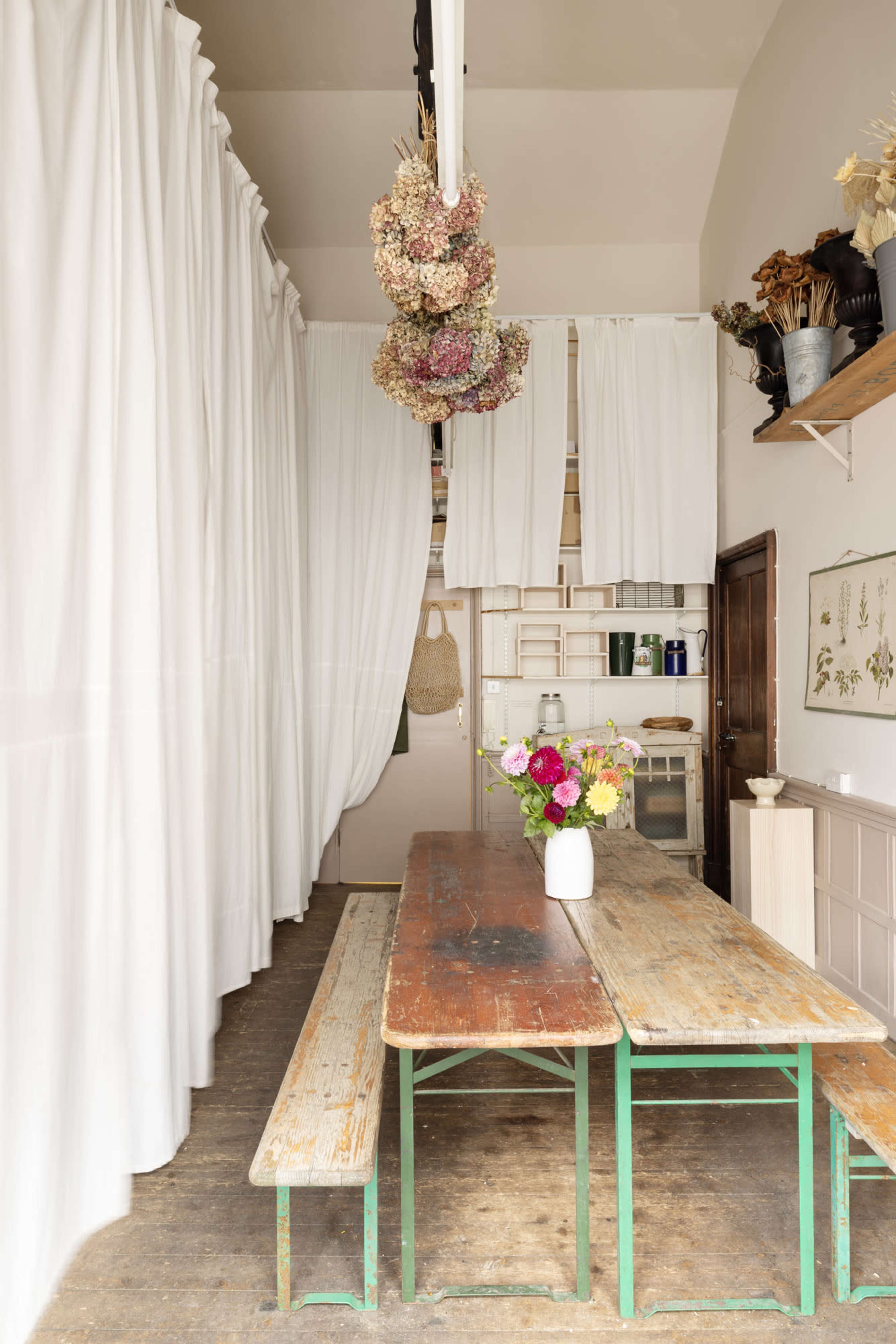 A long, rustic wooden table with benches sits in a bright, airy room adorned with white curtains and dried flowers hanging overhead.