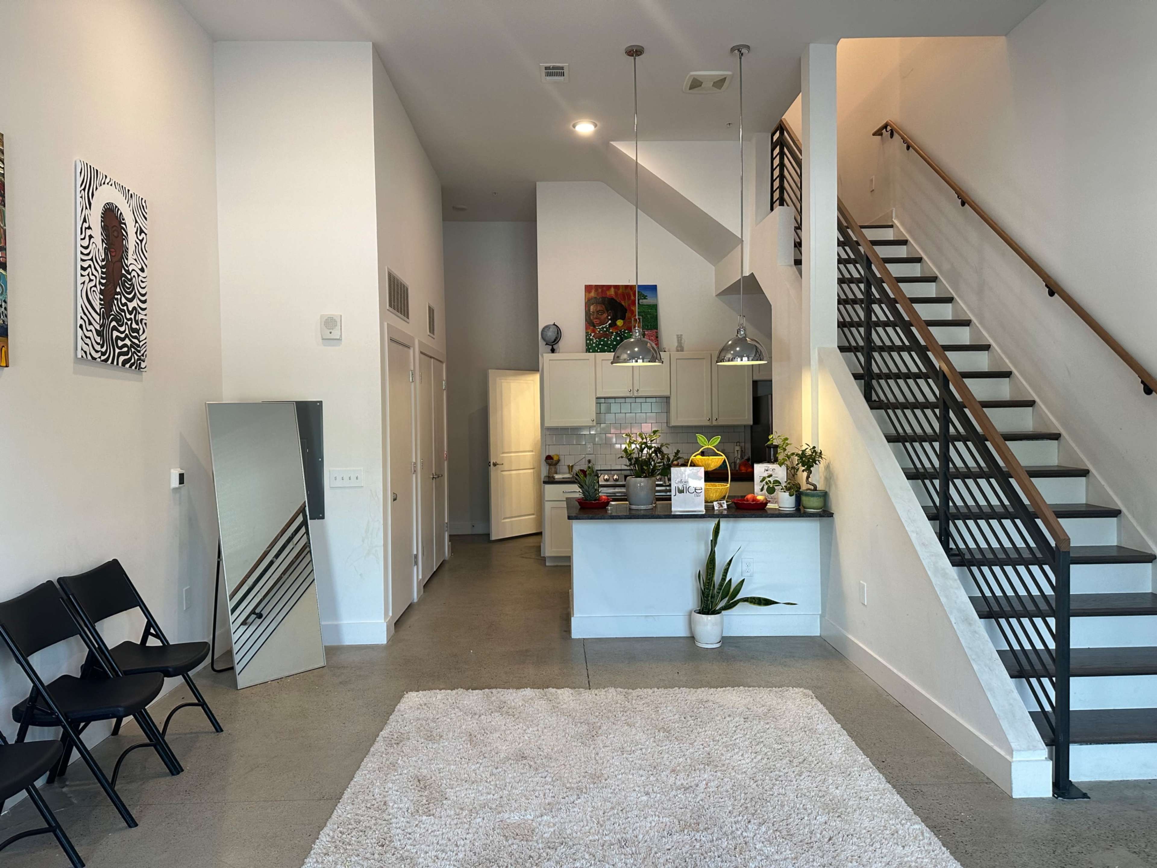 A modern foyer with a staircase, a small kitchen area, a large mirror, plants, and black folding chairs.