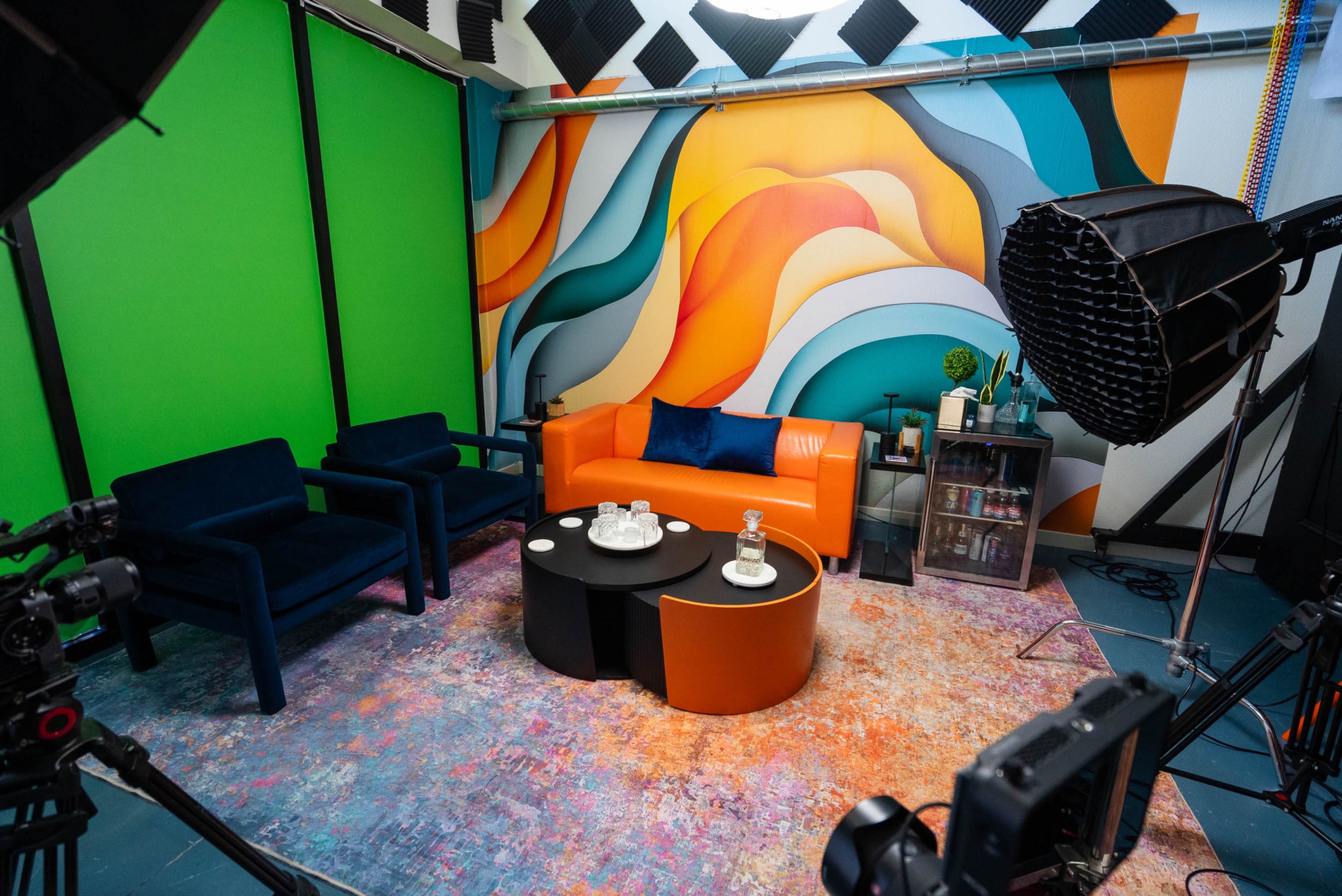 The image shows a colorful studio setup with a vibrant abstract mural, an orange sofa, black and blue chairs, and various filming equipment.