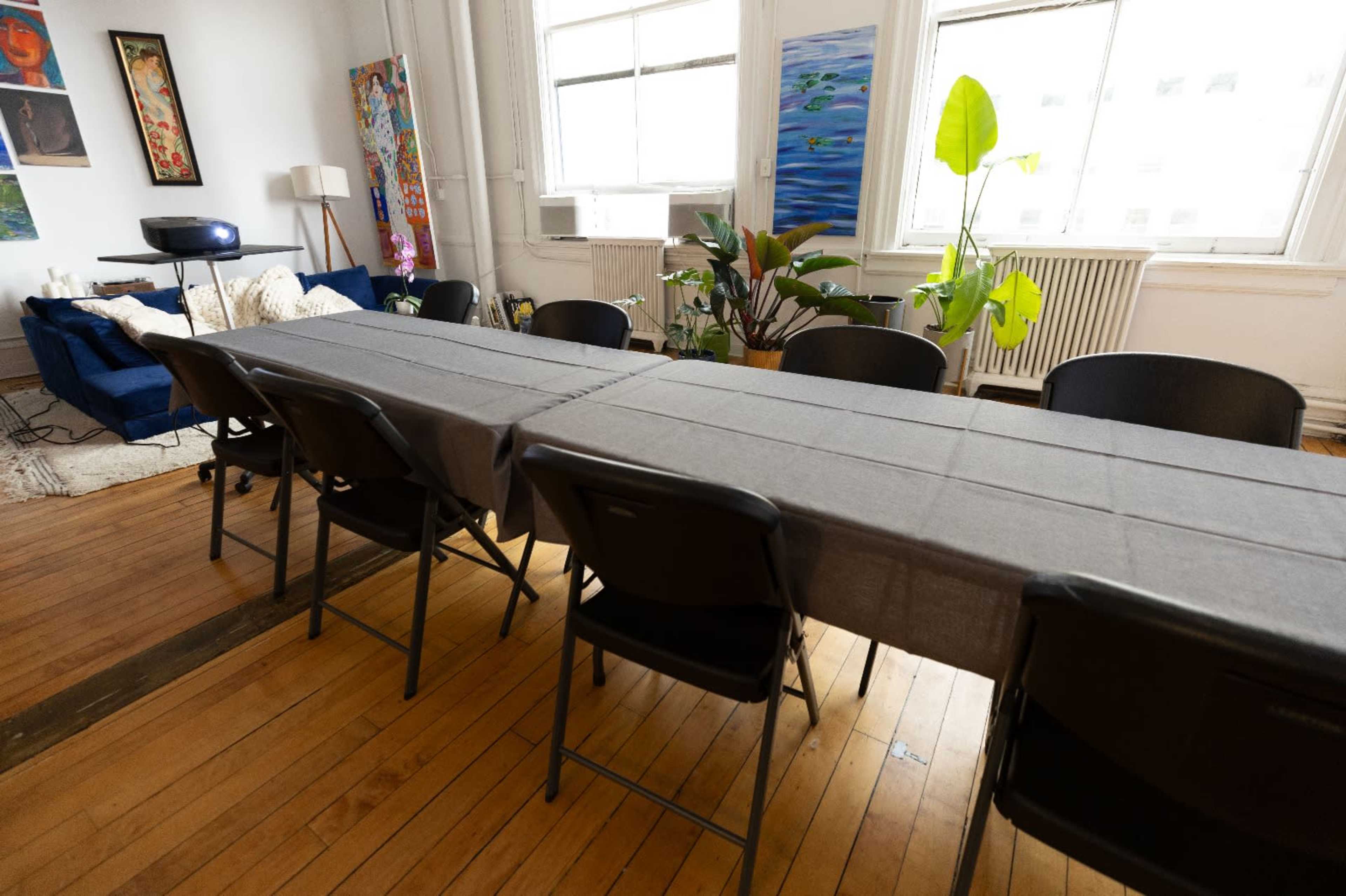 A long, gray table with black chairs is set up in a bright room featuring large windows and potted plants, alongside colorful artwork on the walls.