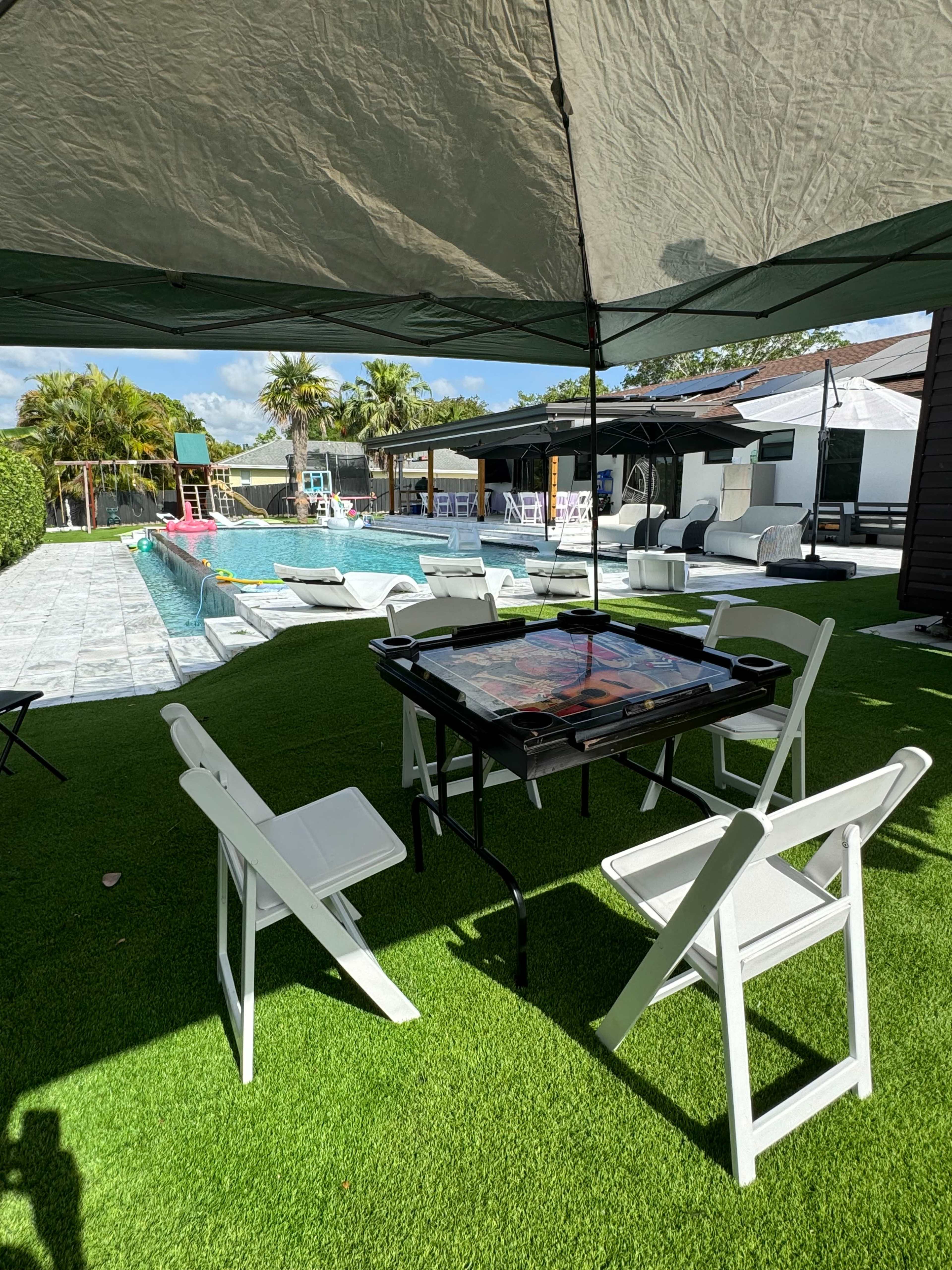 A shaded table with folding chairs is set up on green synthetic grass near a pool surrounded by lounge chairs and palm trees.