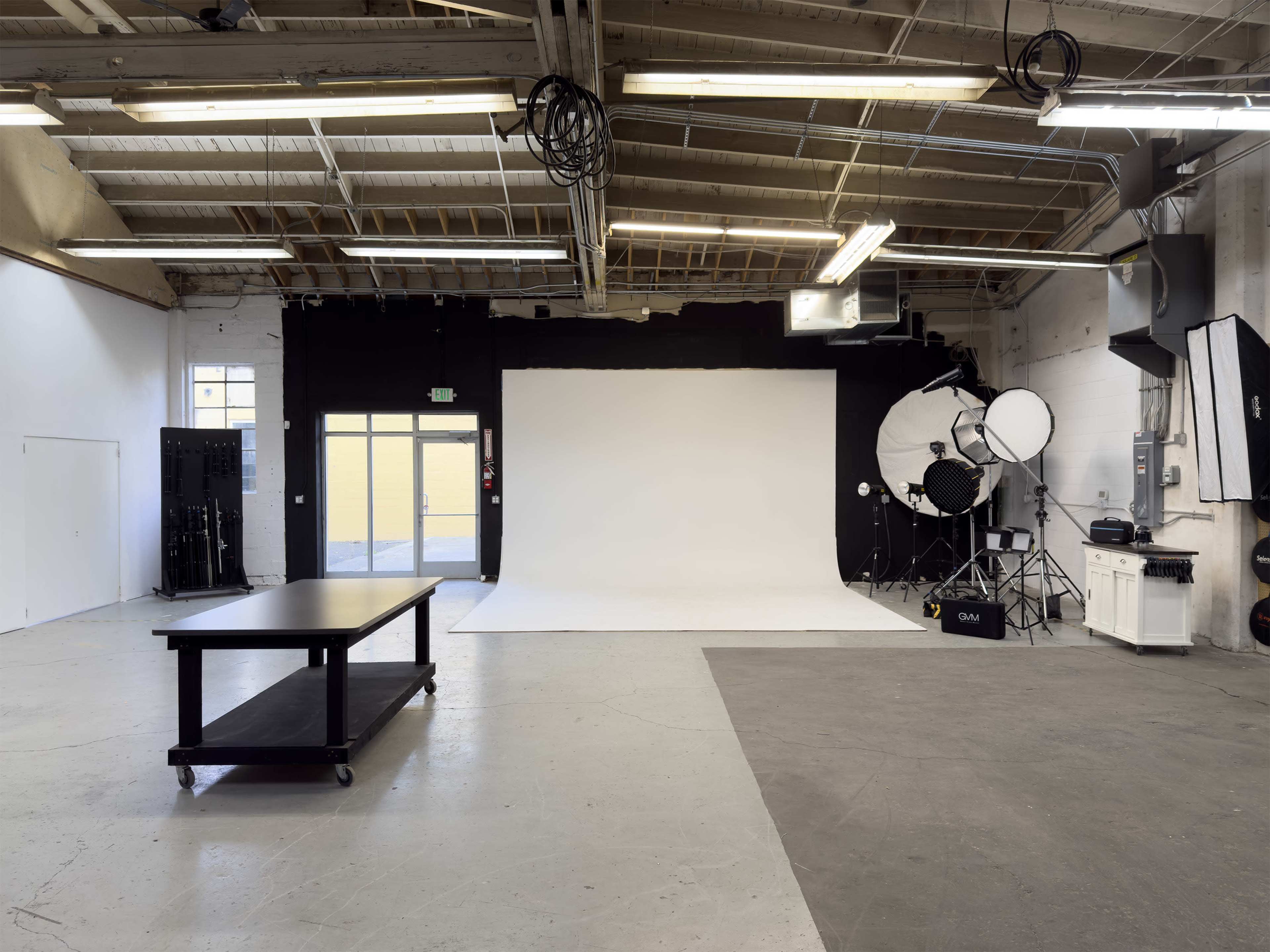 The image shows a spacious photography studio with a large white backdrop, a dark wall, and various lighting equipment arranged around the room.