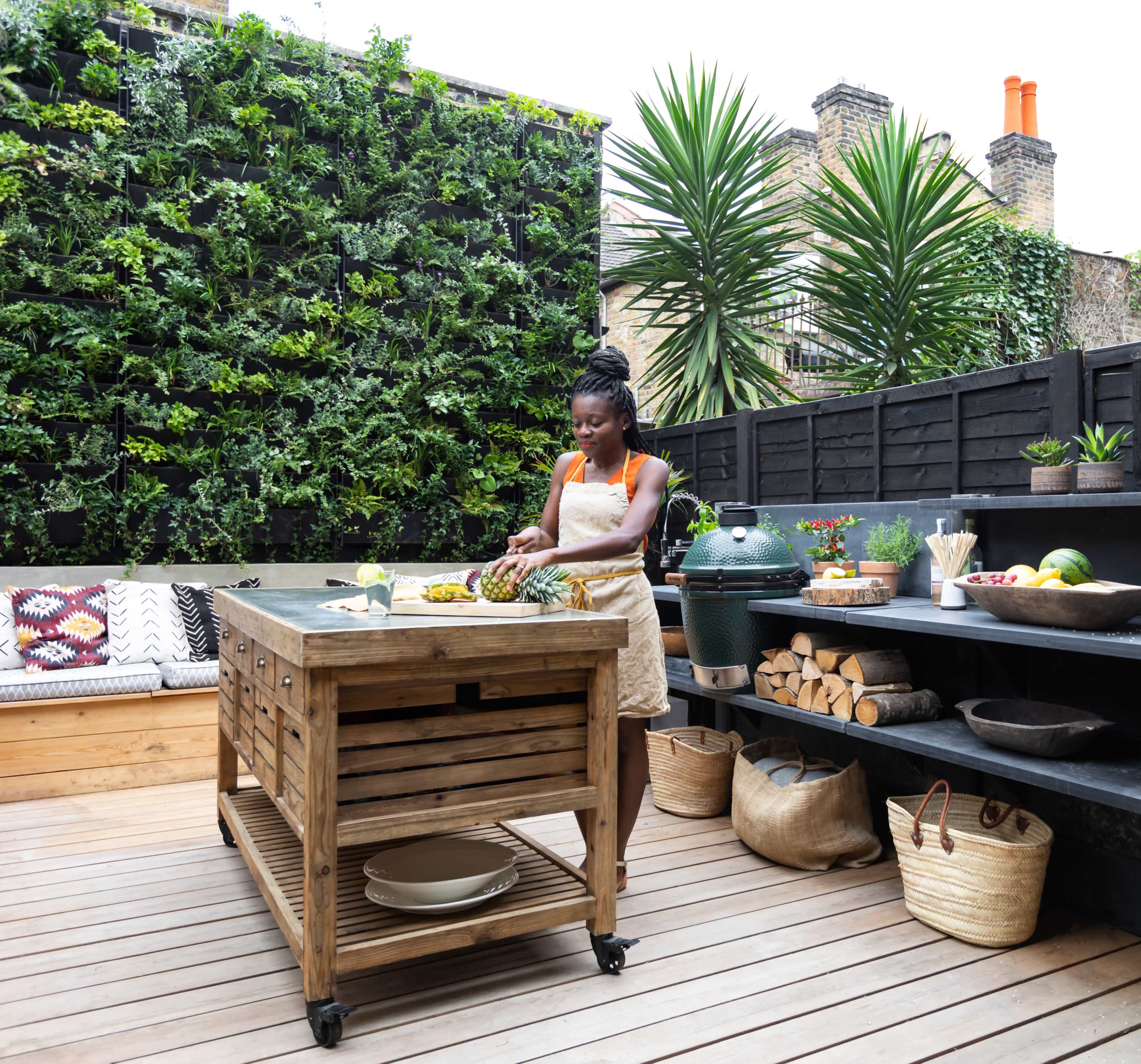 A woman prepares fruit on a wooden kitchen island in a modern outdoor kitchen surrounded by greenery.