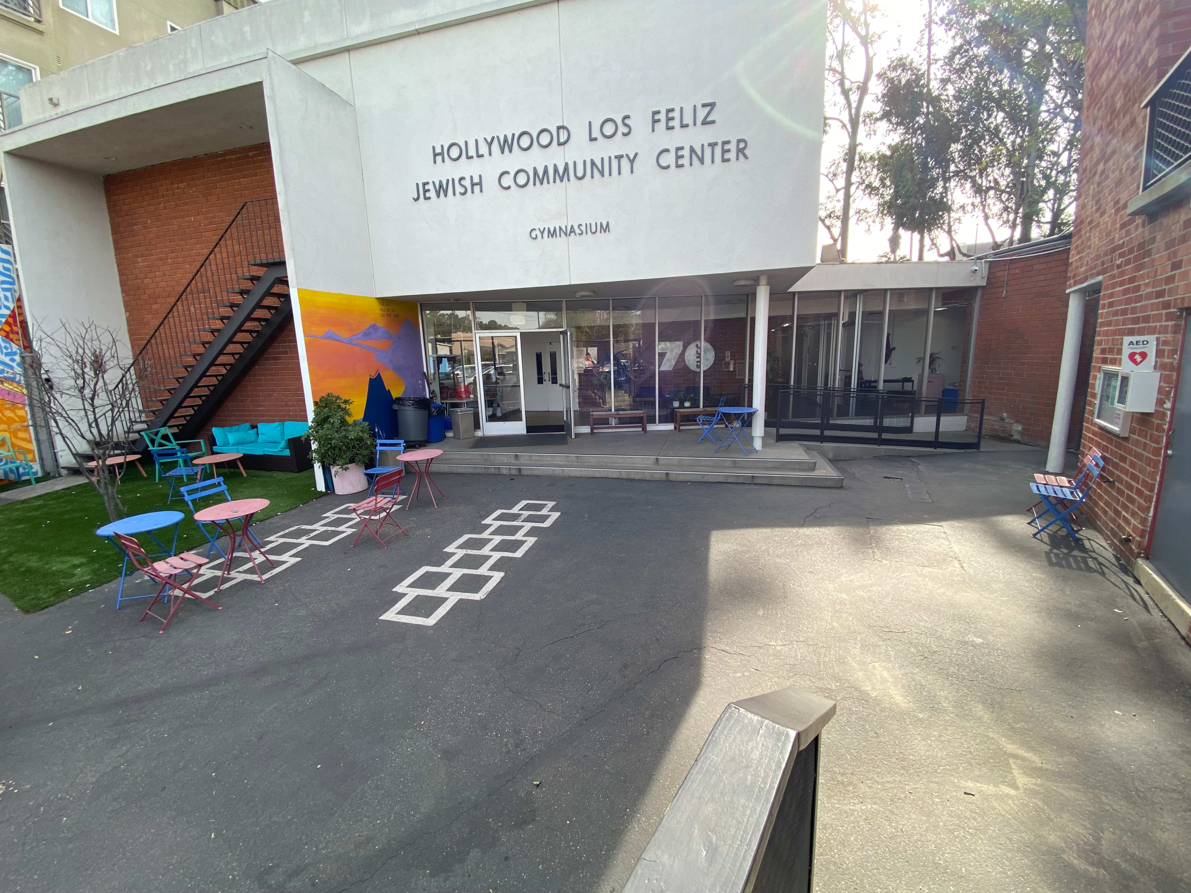 The image shows the entrance to the Hollywood Los Feliz Jewish Community Center, featuring a landscaped area with colorful outdoor seating and a sign indicating the gymnasium.