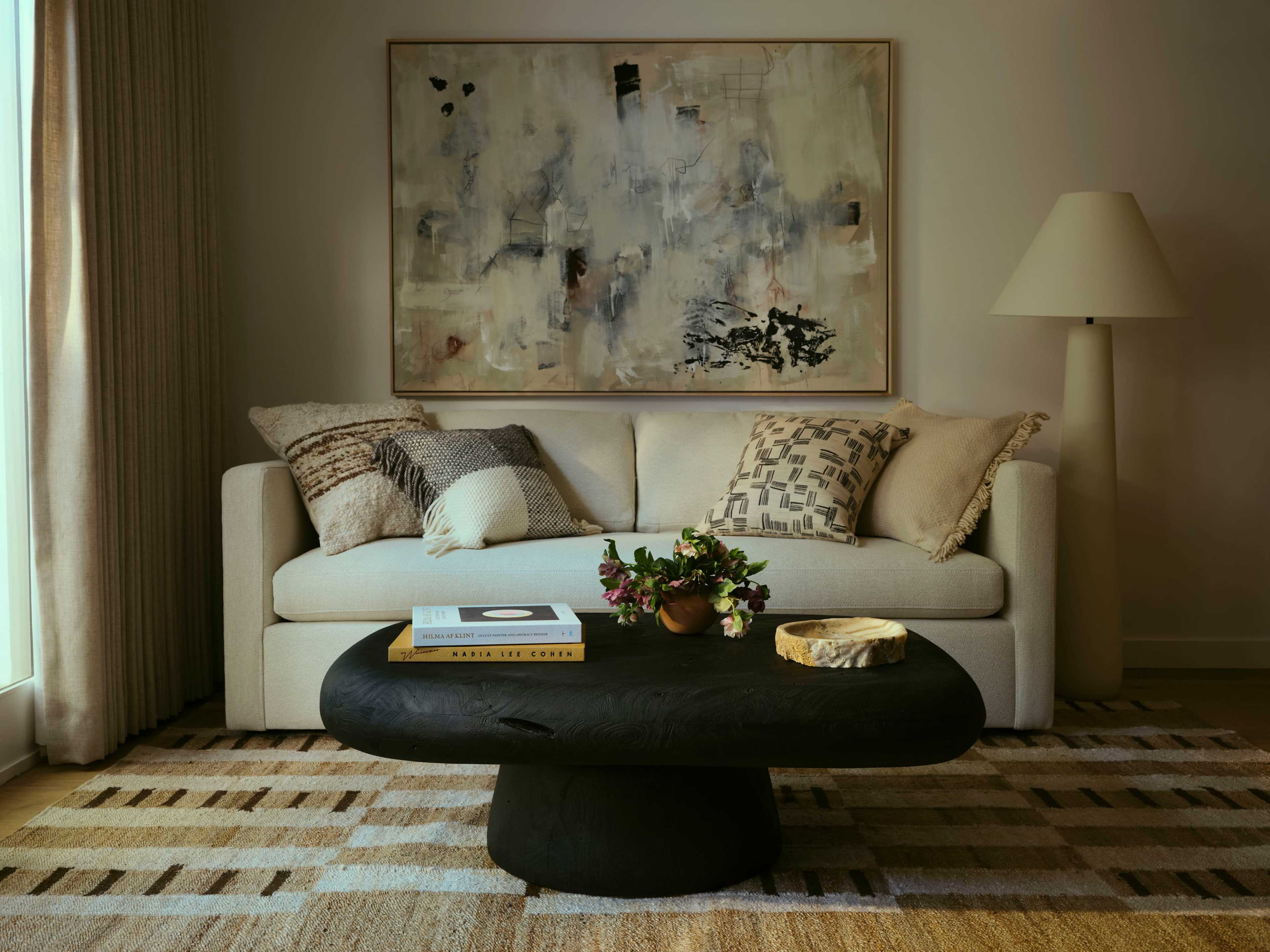 A light-colored sofa with decorative cushions is positioned in front of a round, dark coffee table, under a large wall painting, with a lamp and a small vase of flowers nearby.