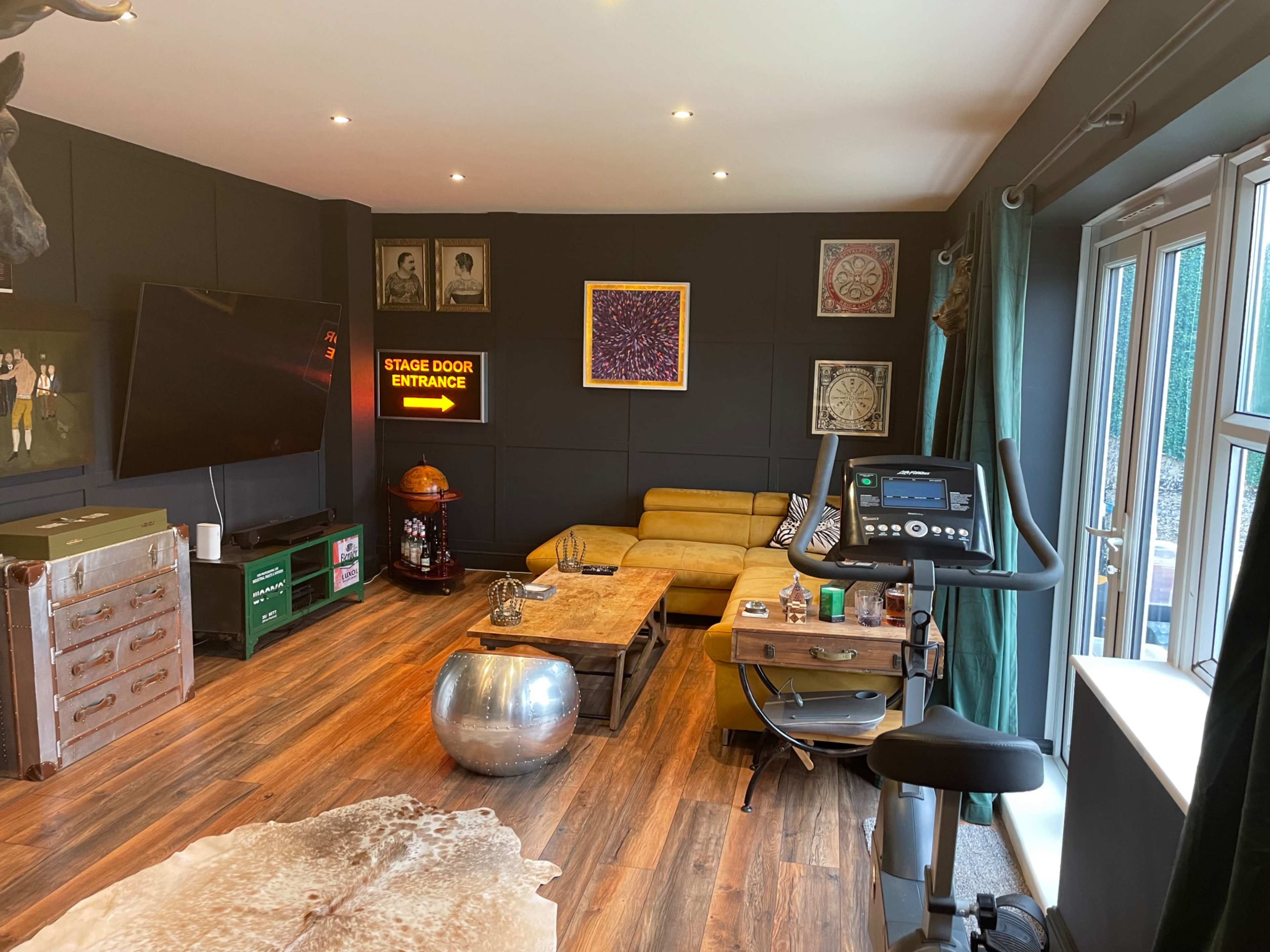 The image shows a modern room with a yellow sectional sofa, exercise bike, and a large television, complemented by various decor items and a wooden floor.
