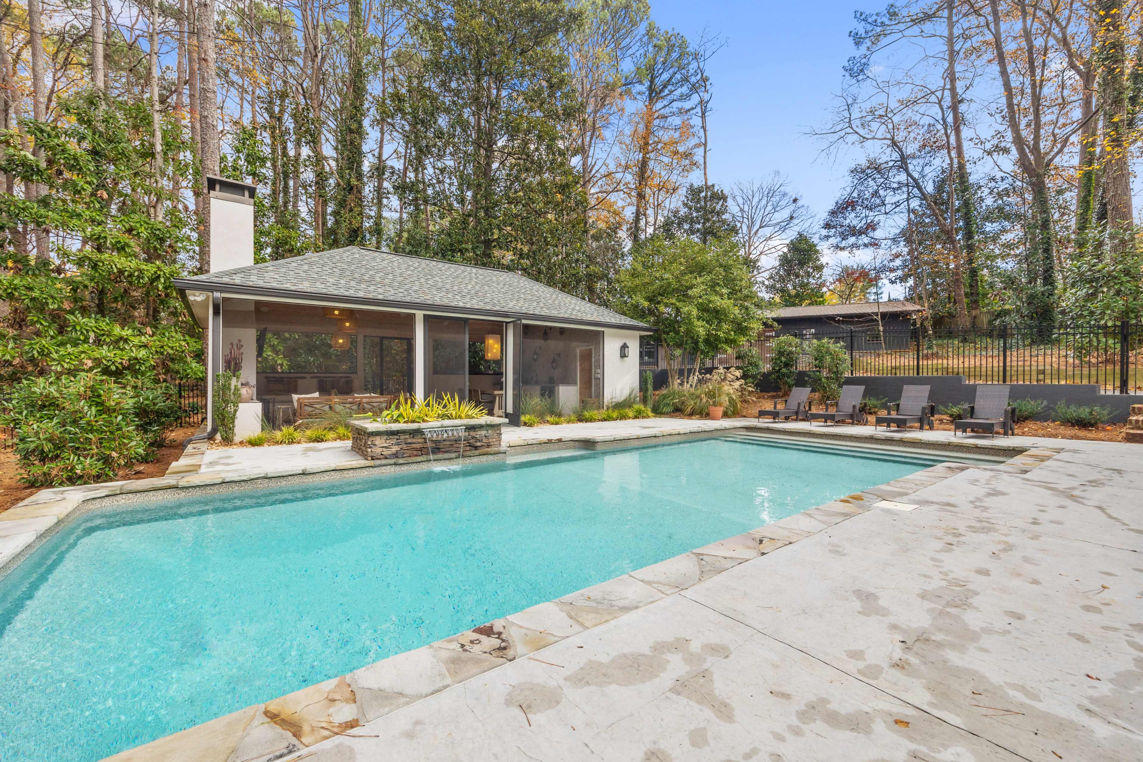 A rectangular swimming pool with a stone border is surrounded by a stone patio and a screened porch, set among trees and landscaping.