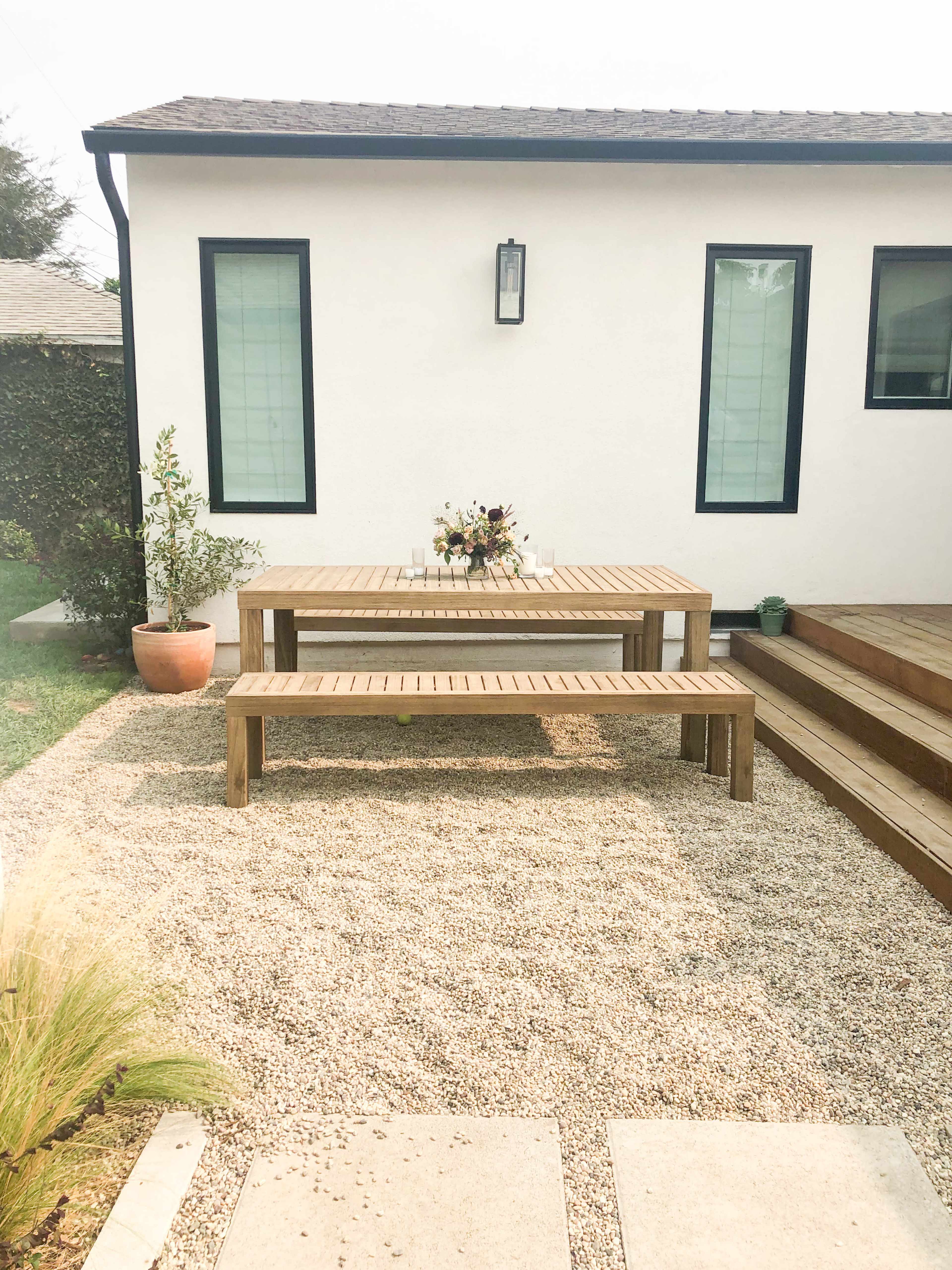 A wooden dining table and benches are placed on a gravel surface next to a white house with large windows.