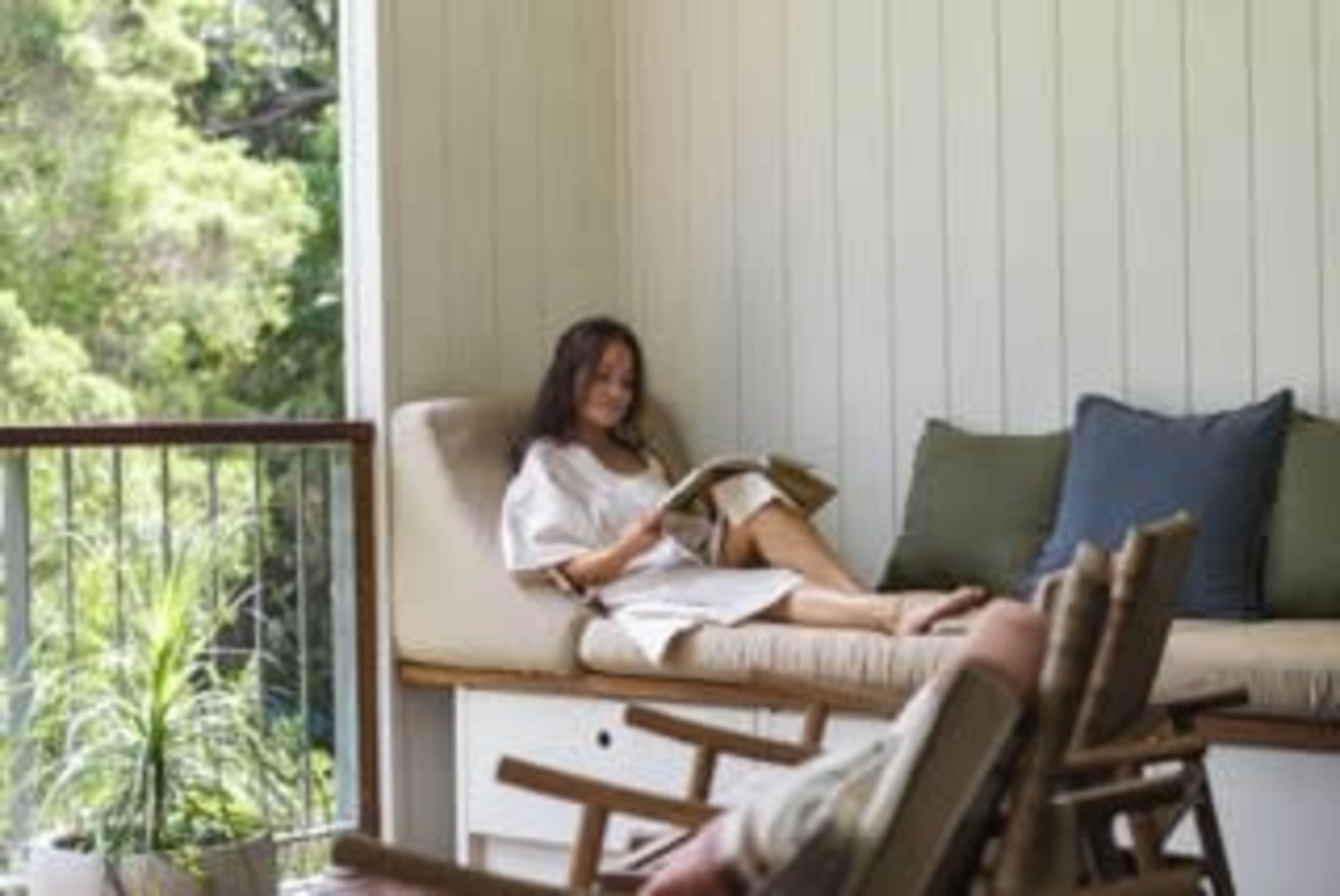 A woman relaxes on a cushioned sofa while reading a magazine in a sunlit, indoor space.