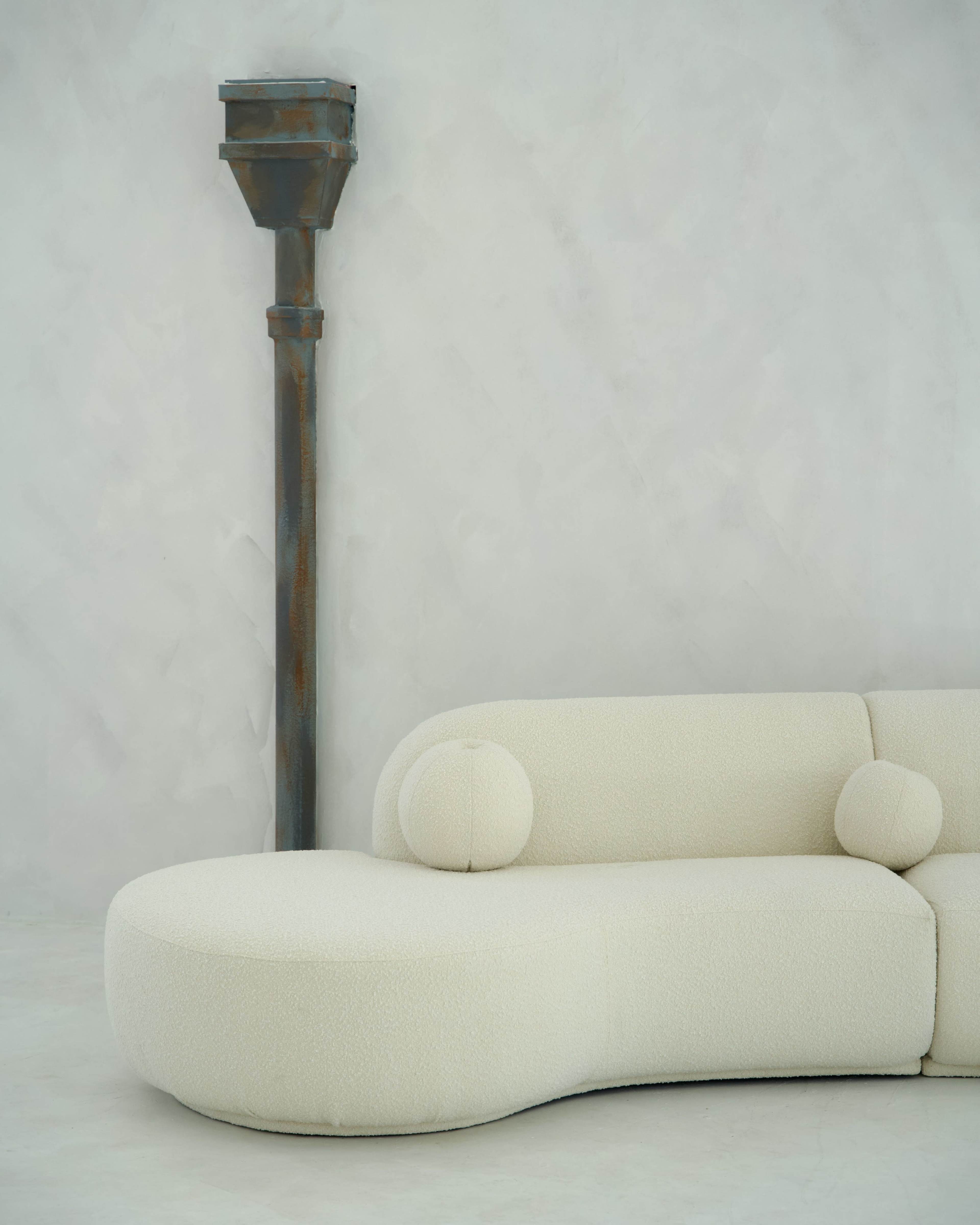 A curved white sofa with round cushions is positioned beside a tall, rust-colored column in a minimalist interior setting.
