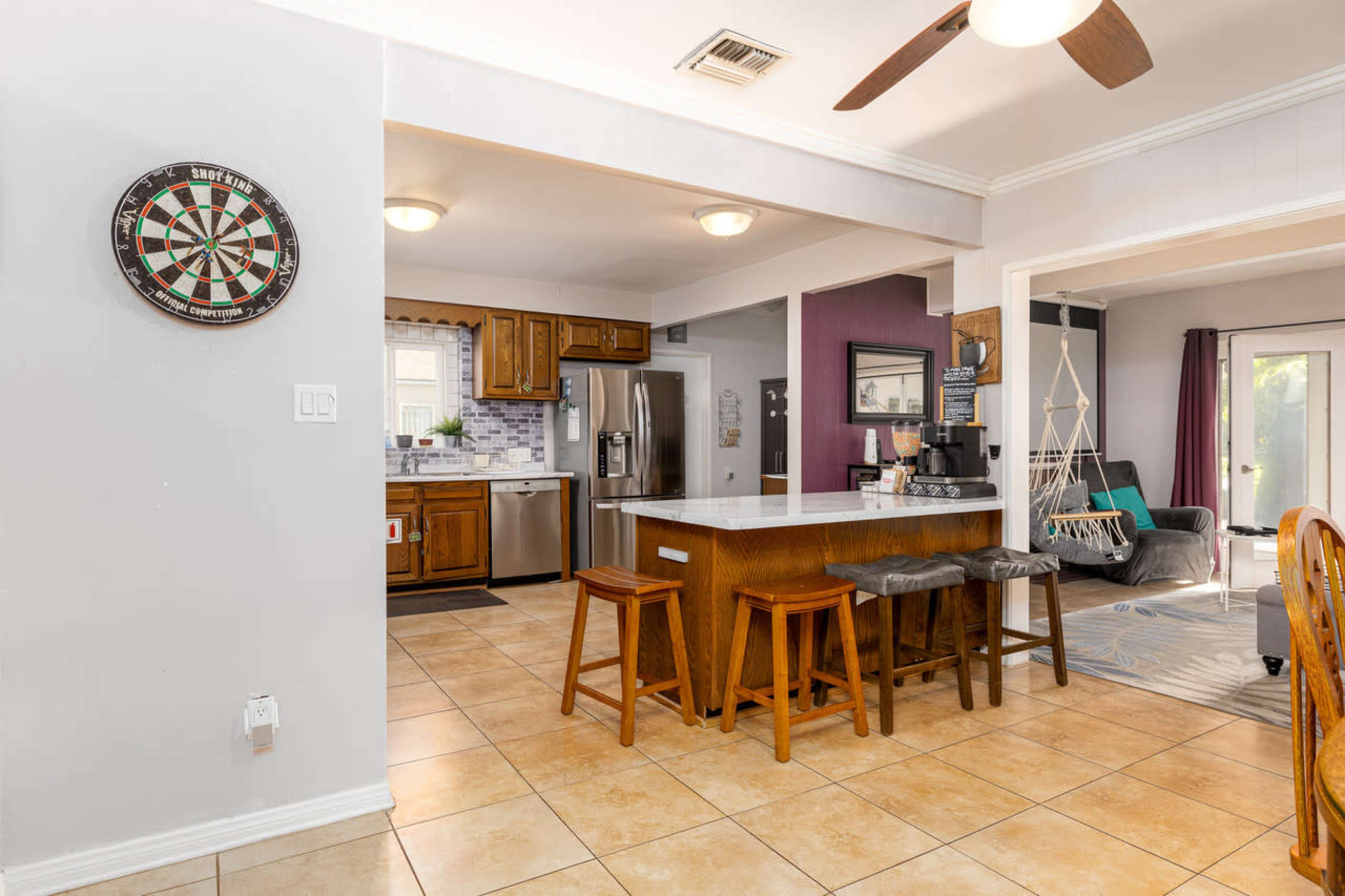 The image shows a spacious kitchen and dining area with wooden cabinetry, a center island with seating, and a dartboard mounted on the wall.