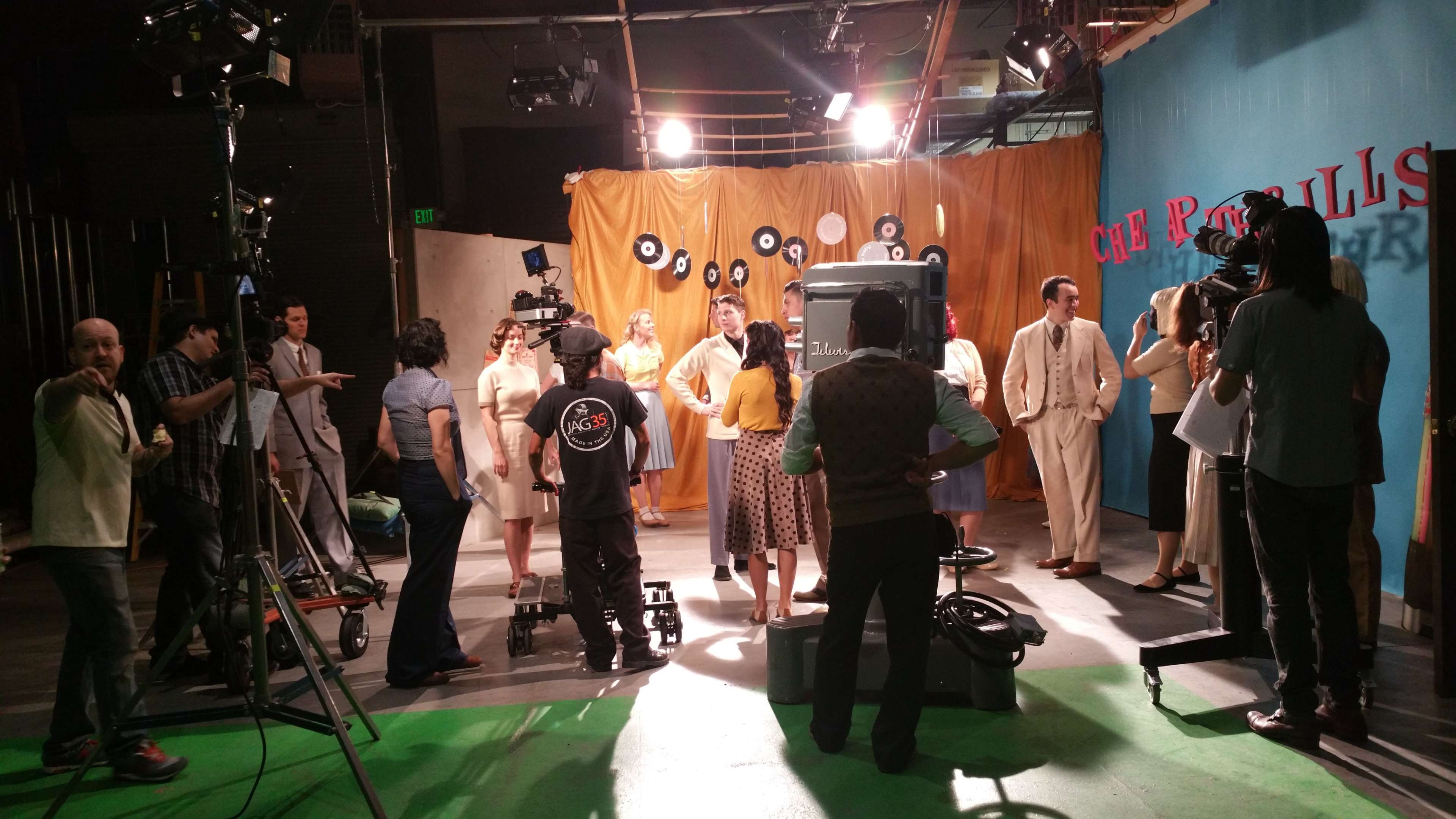 A cast and crew are gathered on a television set with vintage decor and equipment, preparing for a shoot.