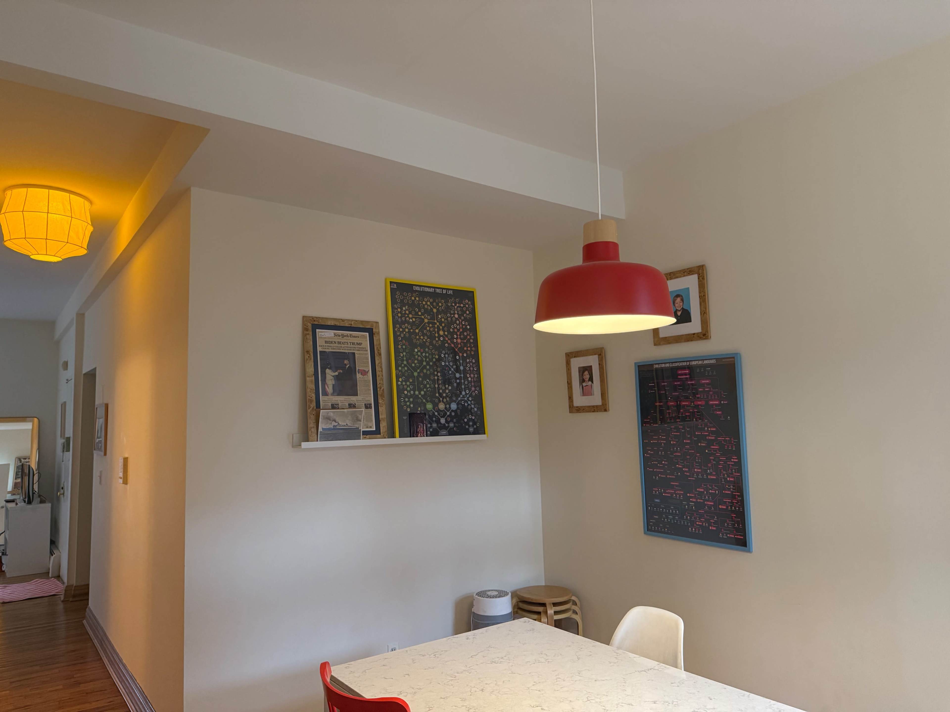 The image shows a dining area with a table, a red pendant light, and framed artwork on the walls.