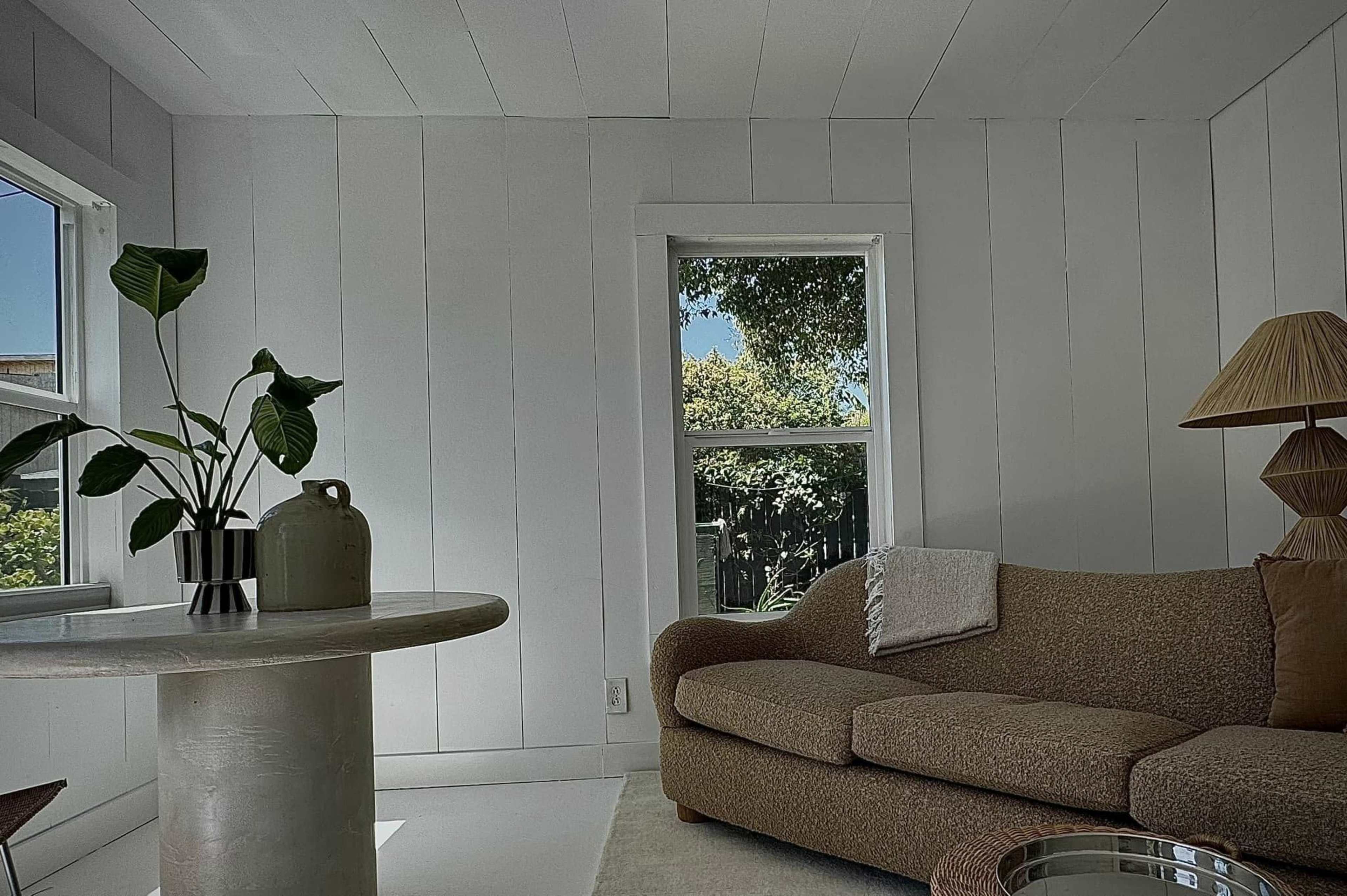 The image shows a minimalistic living room with a beige sofa, a round table, a decorative lamp, and a potted plant by the window.