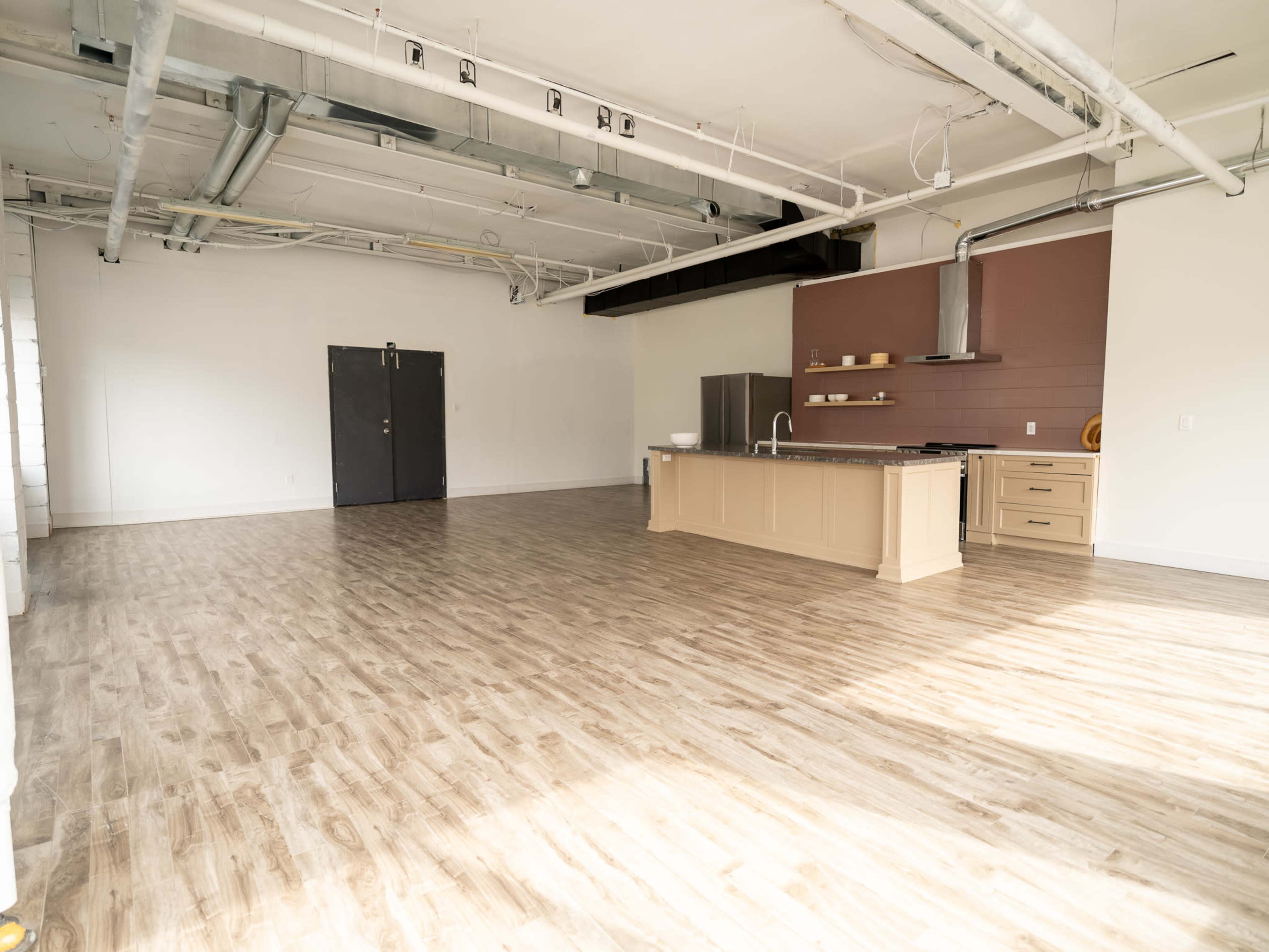 The image shows a spacious, empty kitchen area with wooden flooring, a central island, and modern appliances against a plain wall.