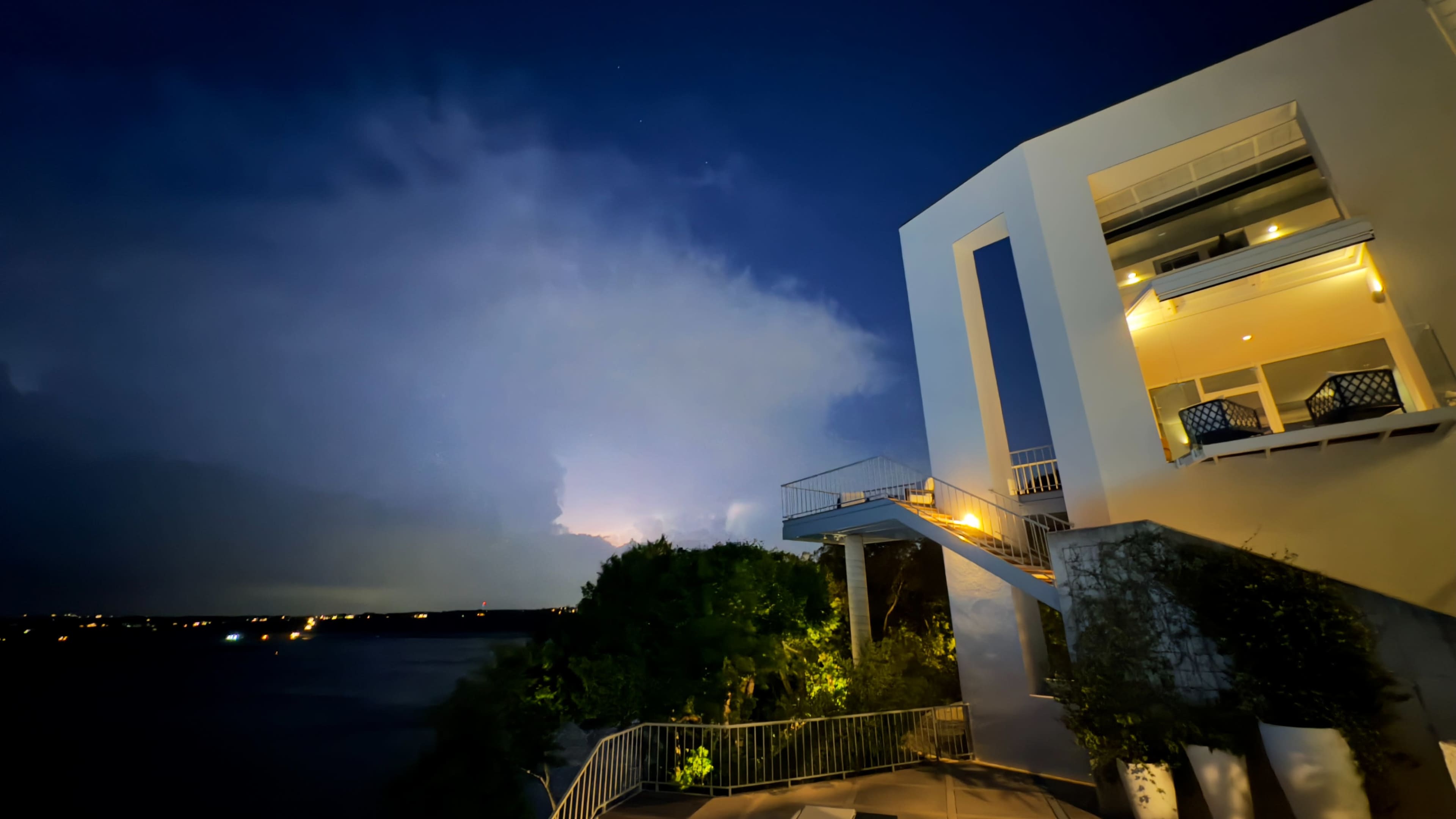 A modern building overlooks a body of water under a darkening sky illuminated by distant lightning.
