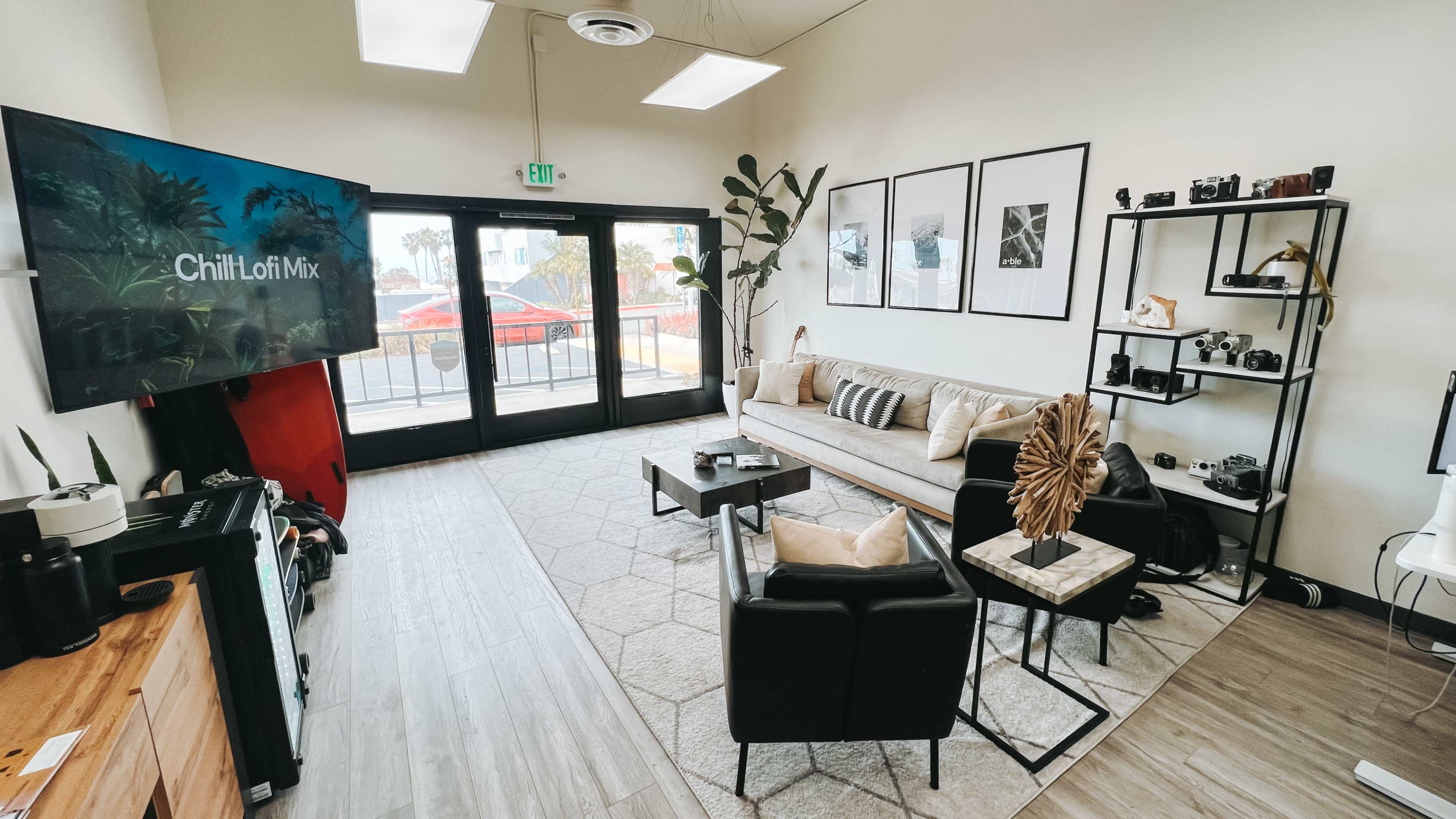 The room features a modern living space with a large TV, a beige couch, black chairs, a coffee table, and shelves displaying cameras, against large windows overlooking a street.