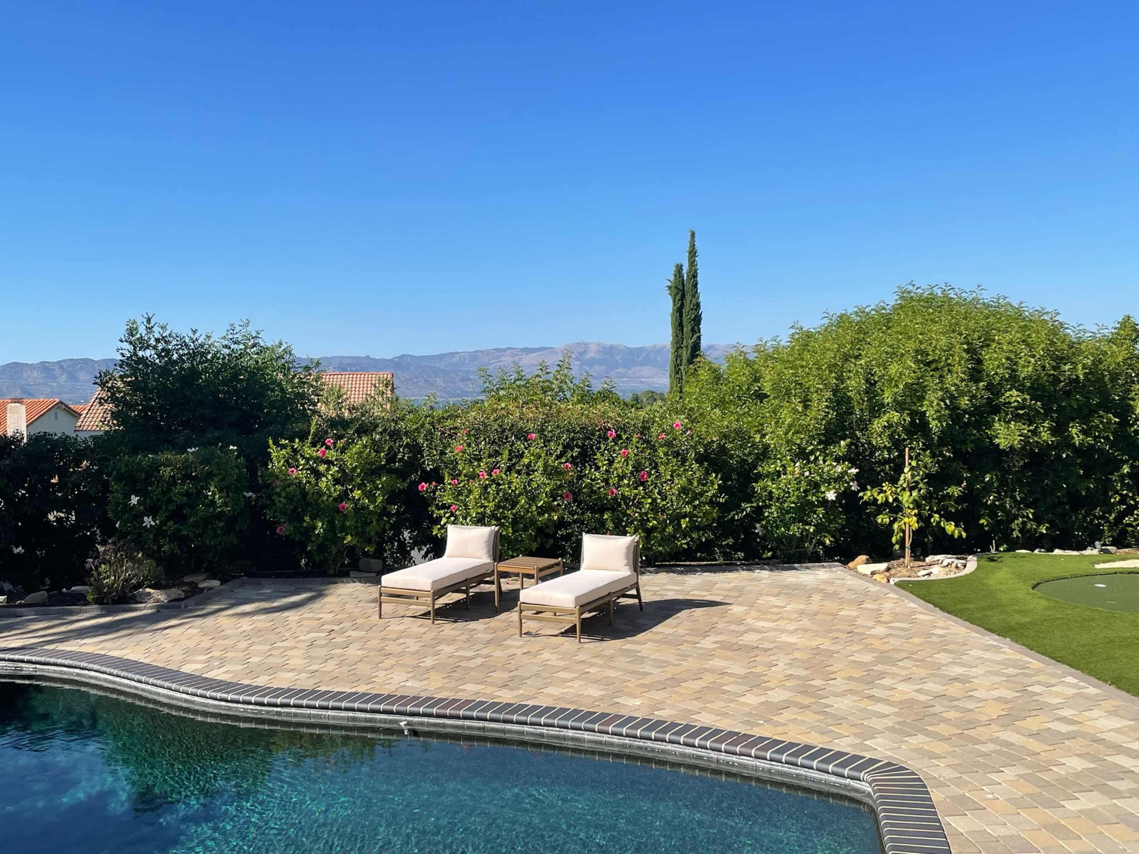 A patio area features two lounge chairs beside a pool, surrounded by green foliage and mountains in the background under a clear blue sky.