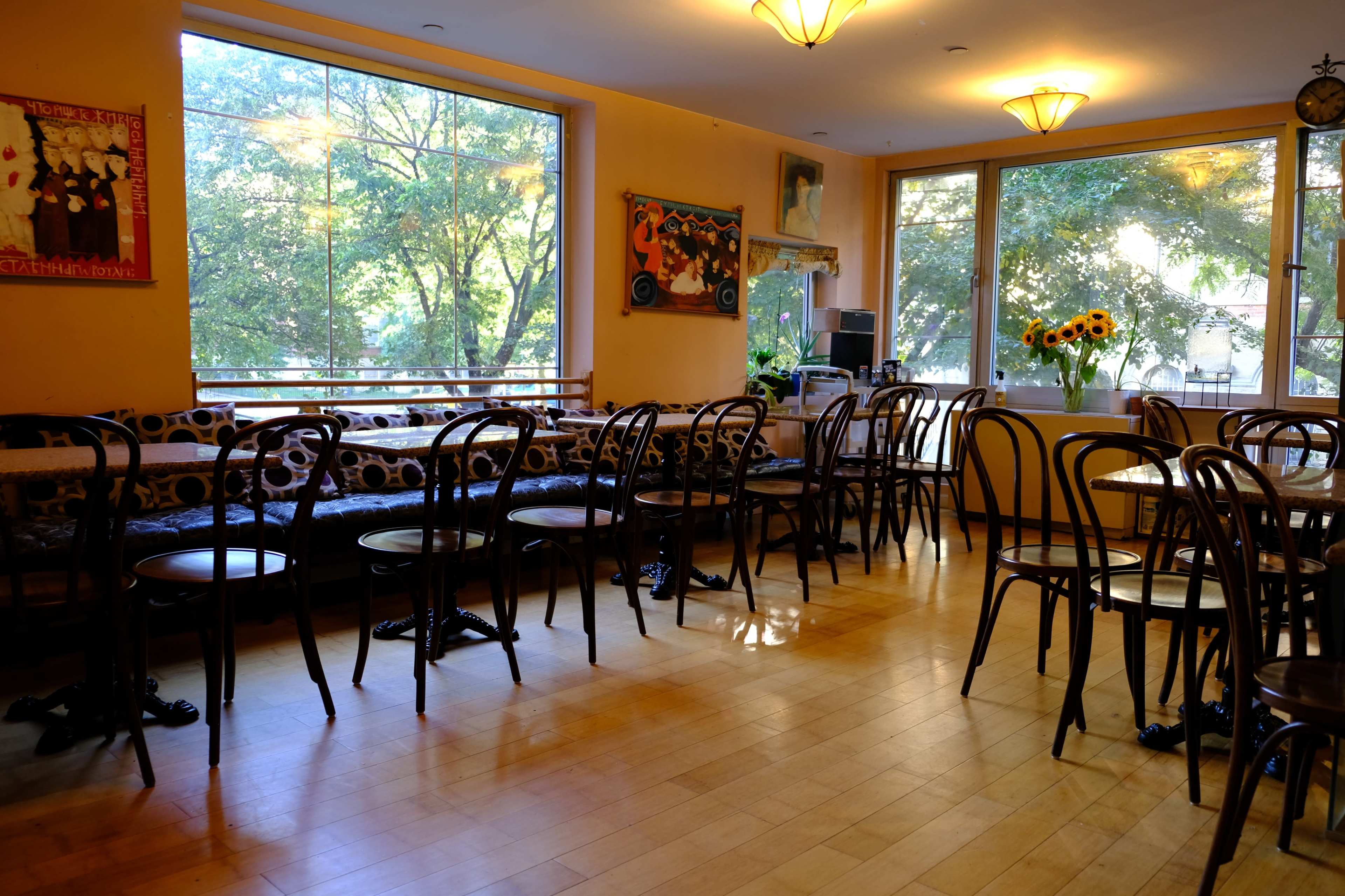 An empty café interior with wooden floors, several chairs arranged at tables, and large windows overlooking trees outside.